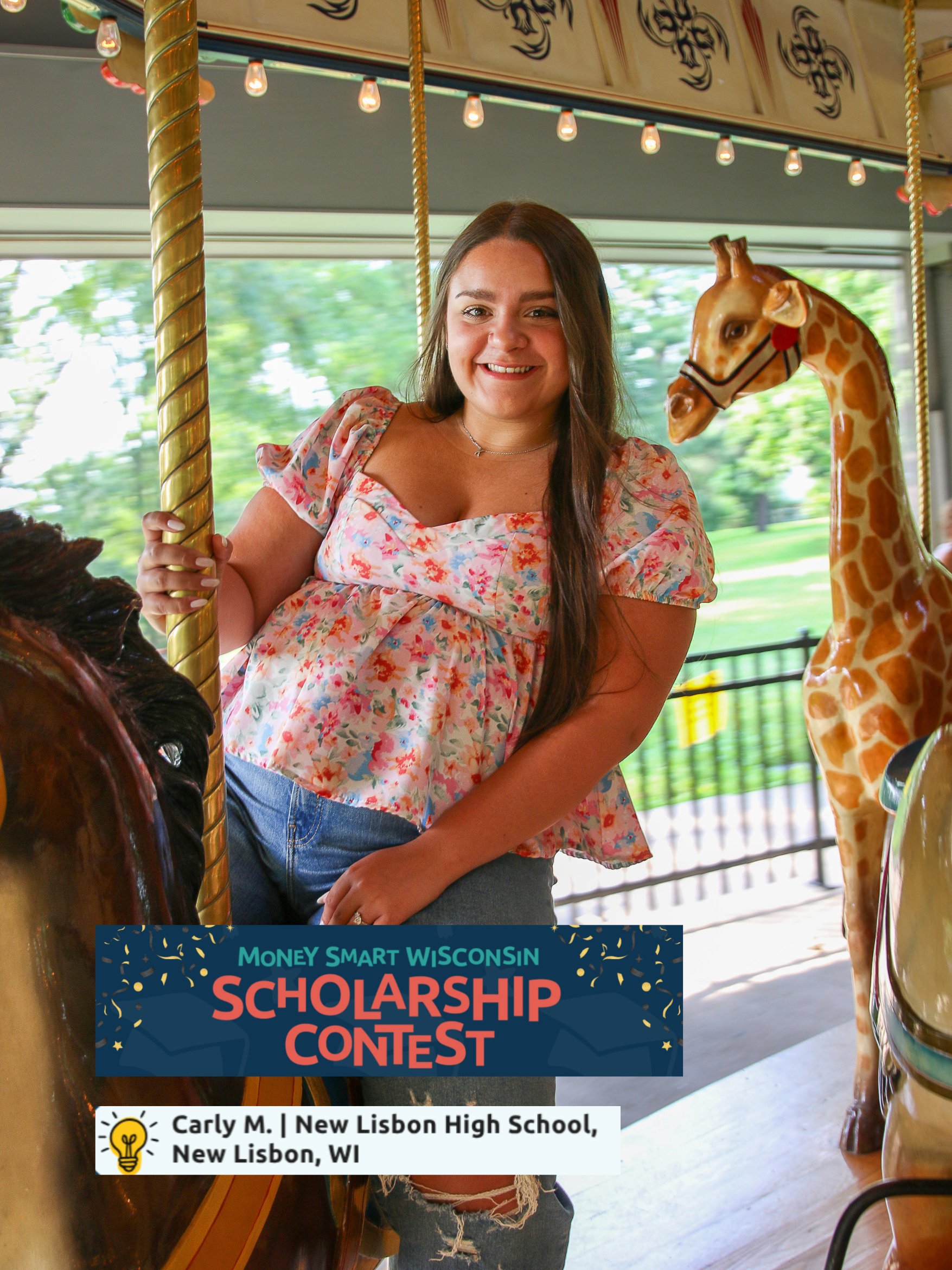 A young woman with long brown hair smiling while riding a carousel horse at an amusement park. She is wearing a floral top and ripped jeans. The carousel has a giraffe figure and decorative lighting, with a background of greenery and park fencing.