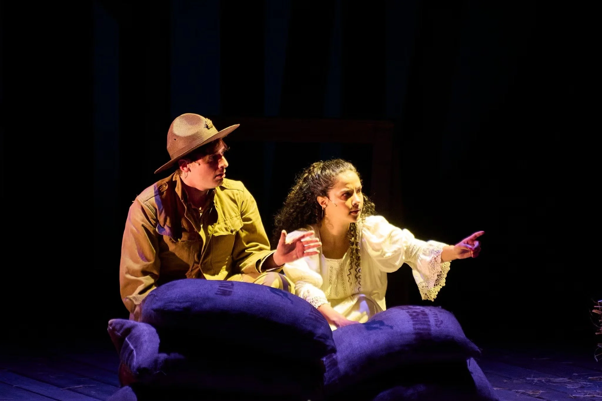 Two actors on stage, a man in a hat and a woman in a white dress, engaged in a dramatic scene, with a dark background.