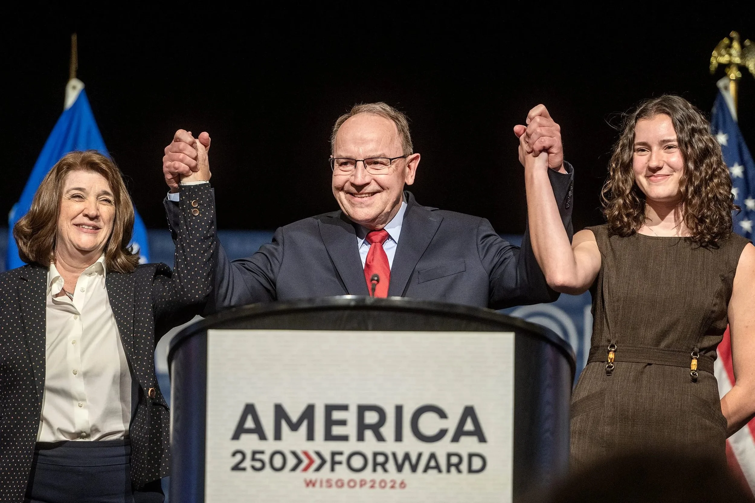 Three people, two women and a man, standing behind a podium with a sign that reads 'AMERICA 250 FORWARD WISGOP 2026'. They are holding hands raised in the air, smiling. The woman on the left has brown hair and is wearing a dark blazer and white shirt. The man in the middle is wearing glasses, a dark suit, white shirt, and a red tie. The woman on the right has curly brown hair and is wearing a brown dress. There are American flags in the background.