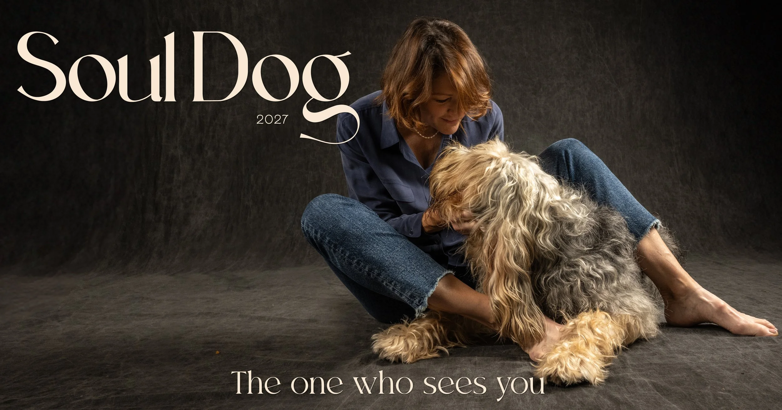 A woman with shoulder-length brown hair sitting on the floor with a curly-haired dog. The woman is smiling and looking down at the dog, which is lying on its back, exposing its belly. The background is dark and textured. The words "SoulDog 2027" are written in large letters in the top left corner, and "The one who sees you" is written at the bottom.