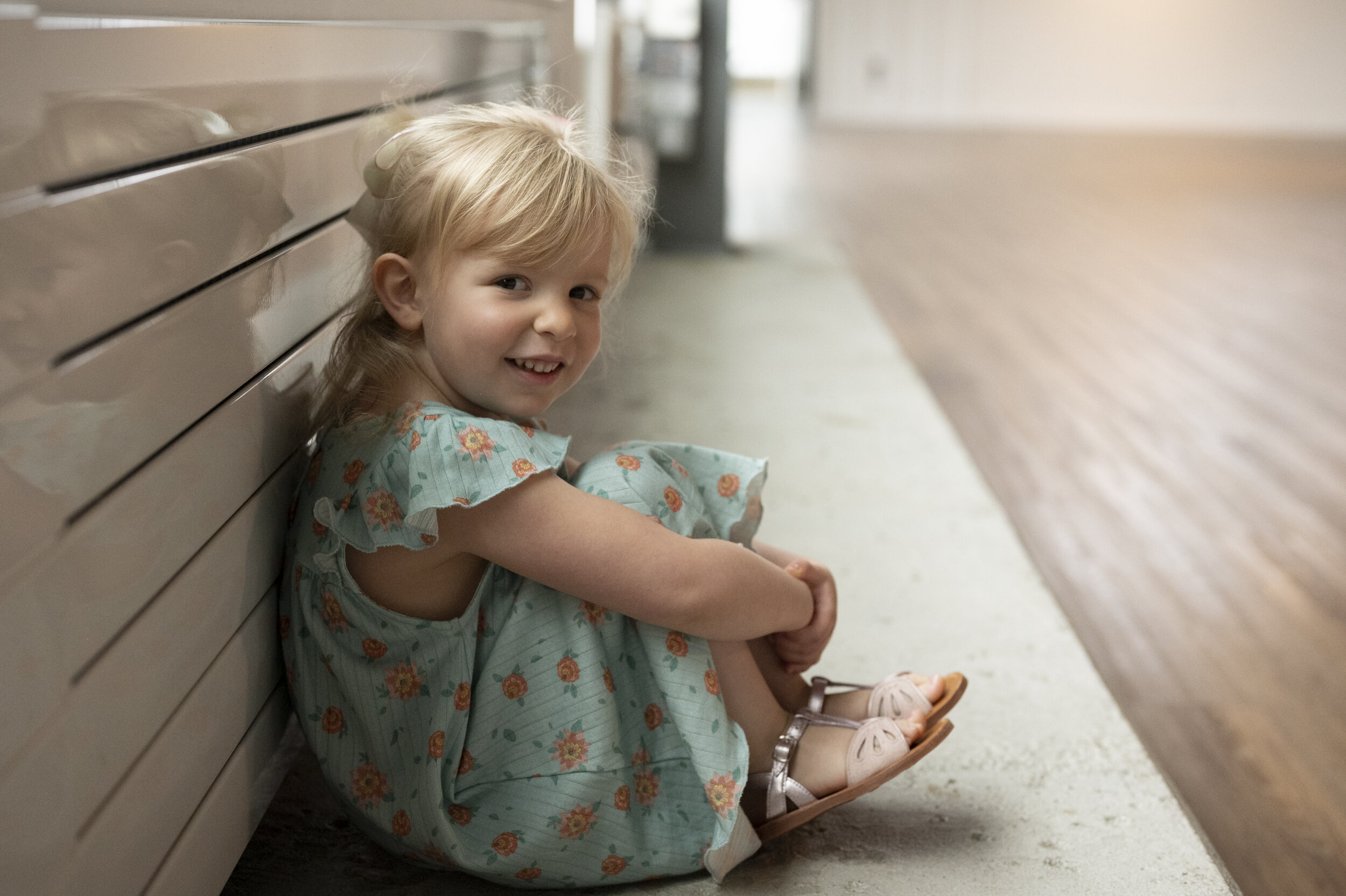 Documentary style photo of a young blond child in a blue dress. She has her back on the wall and is sitting on the floor.