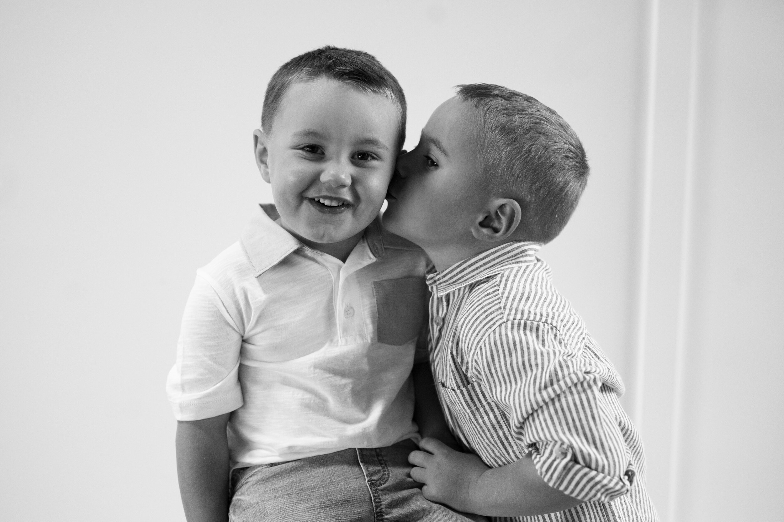 Black and white photo of two boys in white t-shirts. One is kissing the other on the cheek.