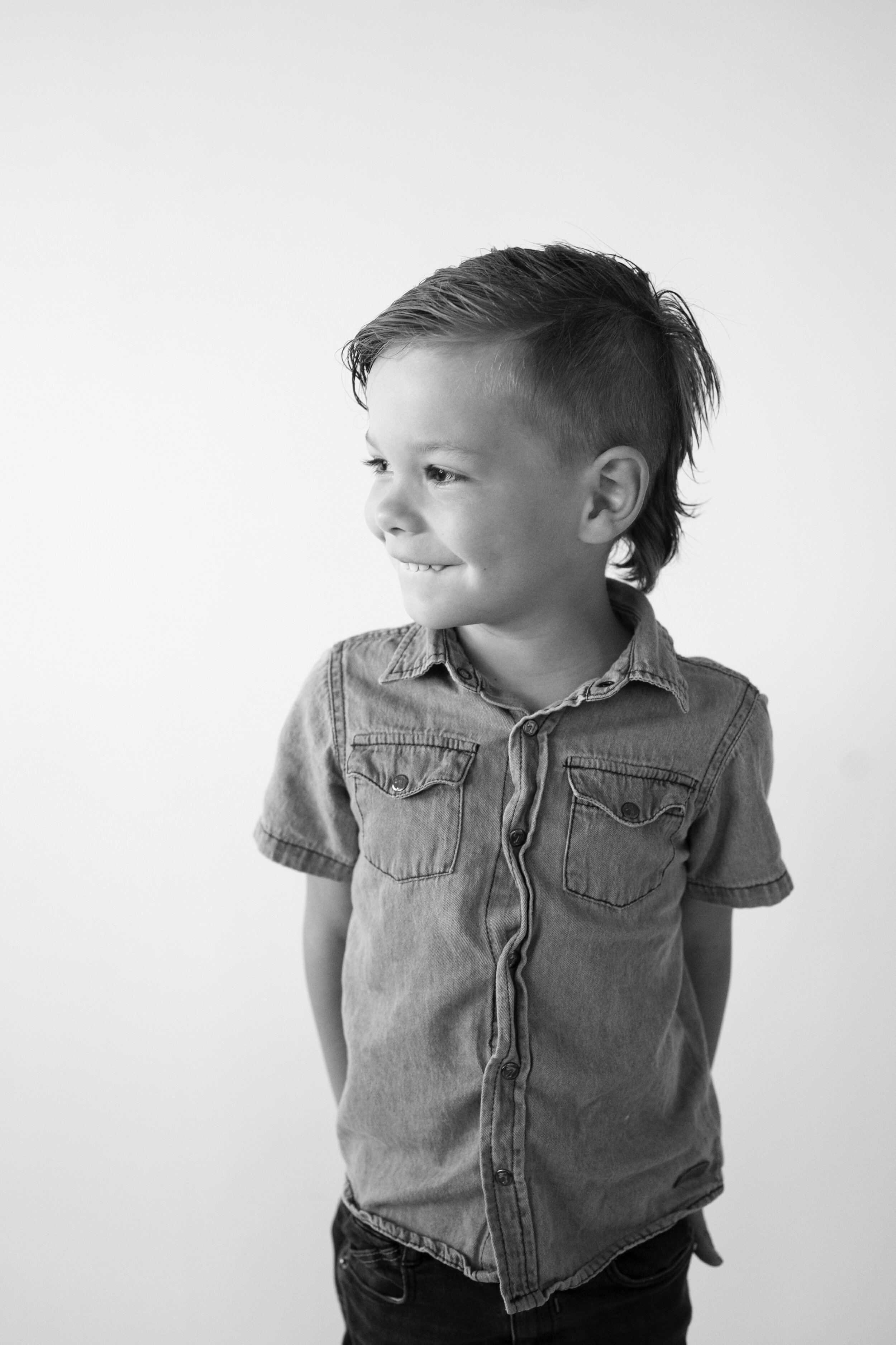 A young boy with a mohawk style hair cut wearing a jean shirt. He is smiling and looking to the side. He is standing against a white background.