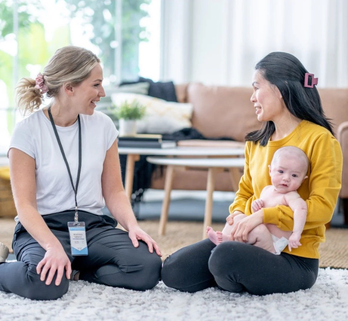 A woman with dark hair and a yellow shirt holding a baby, sitting on a carpet, talking to a smiling woman with blond hair tied with a pink hair tie, sitting on her knees in a bright room with large windows.