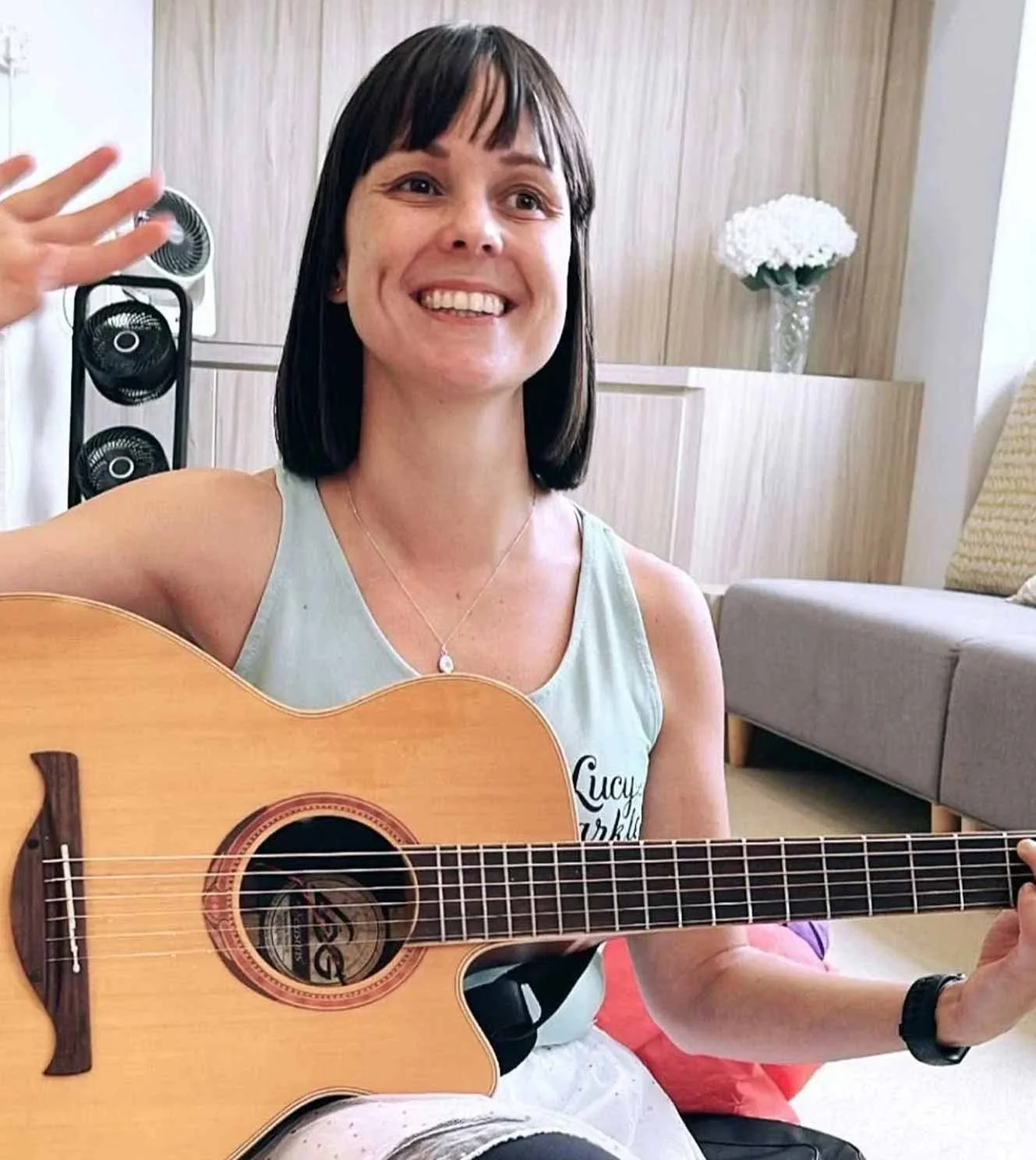 A woman playing an acoustic guitar, smiling, in a room with a wooden panel and flower vase in the background.