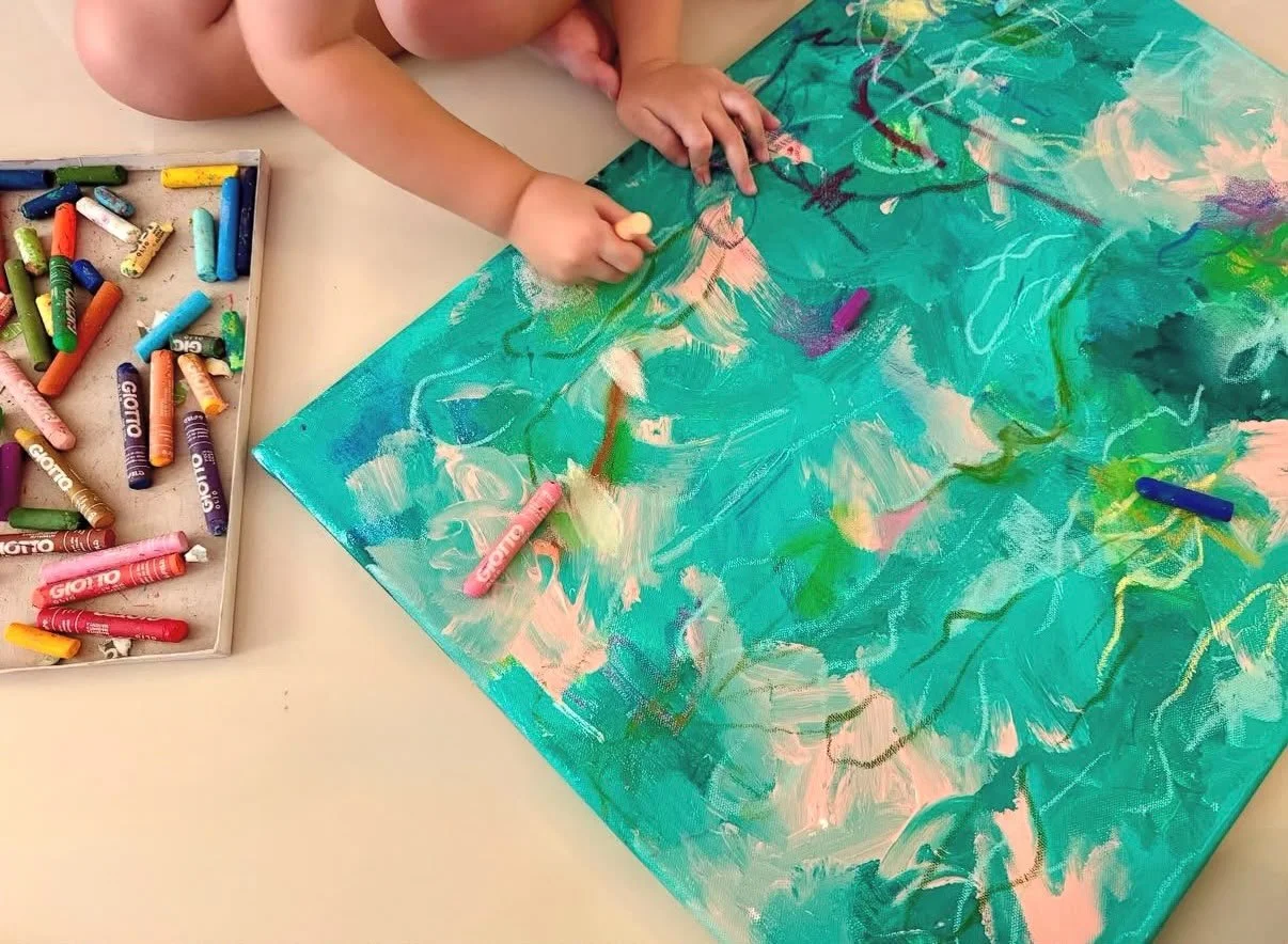 A child is painting on a large canvas using colorful chalks and crayons, with a tray of multicolored chalks nearby.