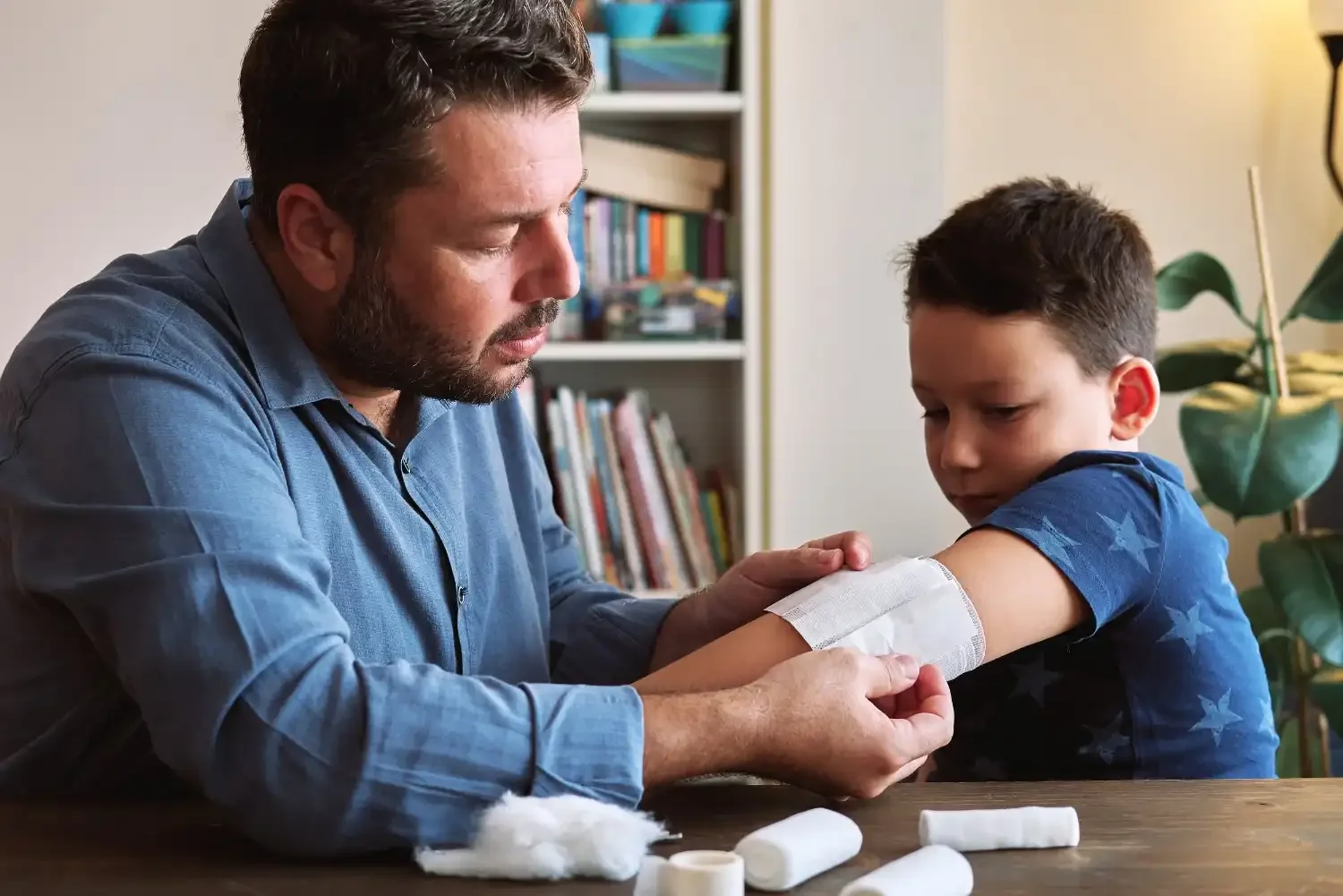 A man is helping a young boy with getting a vaccination shot in his arm. The man appears focused and caring, while the boy is looking down. There are medical supplies on the table, and a bookshelf and houseplant are visible in the background.