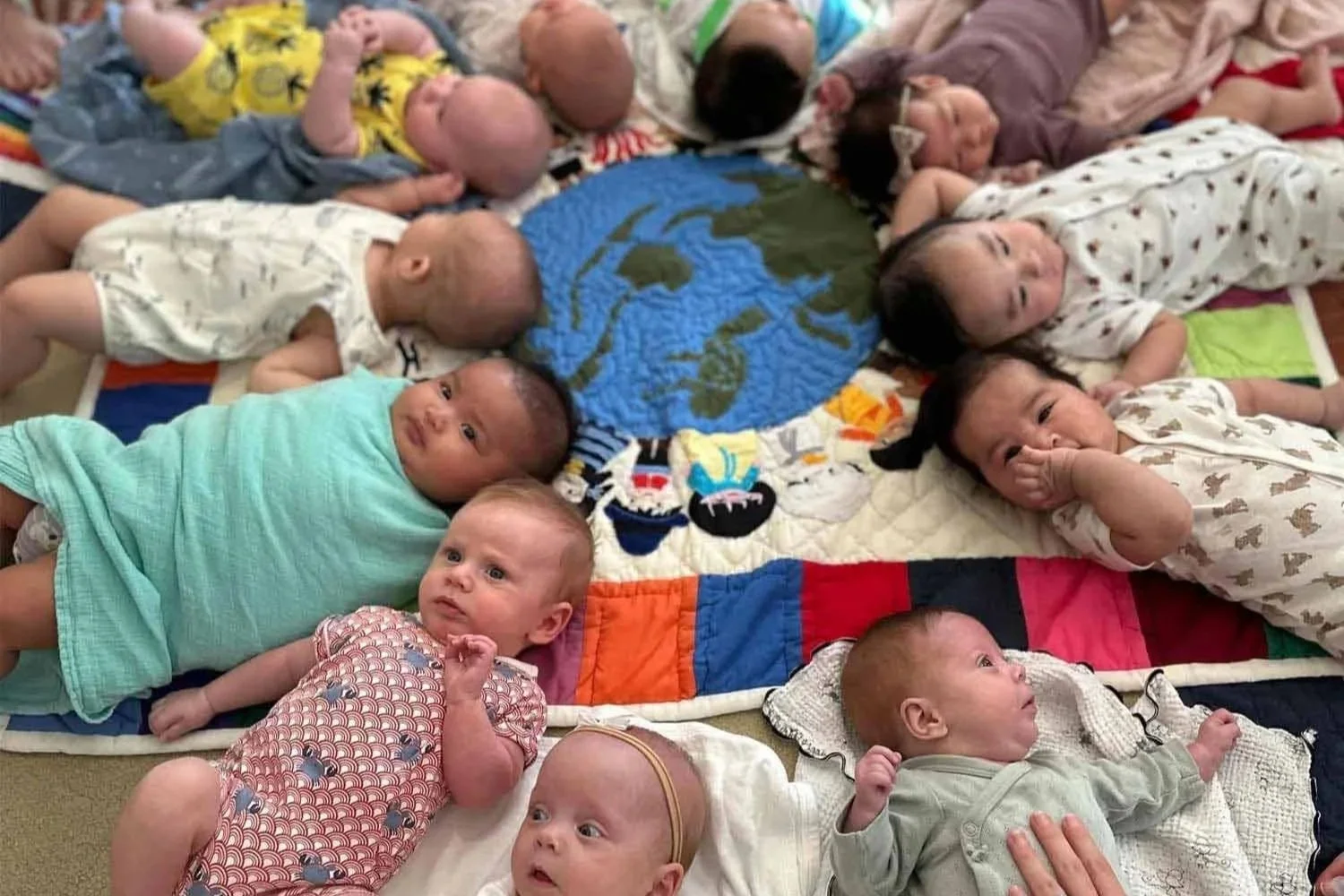 Group of 11 babies lying on their backs on a colorful quilt with a world map design, some looking at the camera, others resting or looking around.