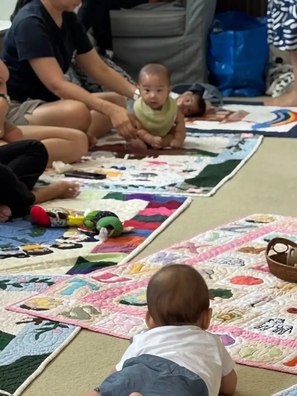 Children and adults sitting and lying on colorful quilted mats on the floor, with some toys nearby; an indoor setting.
