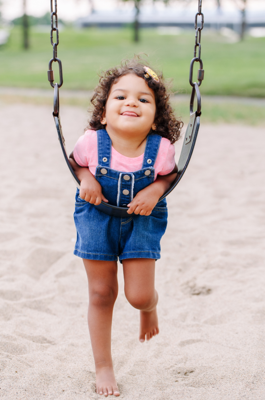 Young girl on a swing at the playground, smiling and sticking out her tongue, wearing a pink shirt and denim overalls.