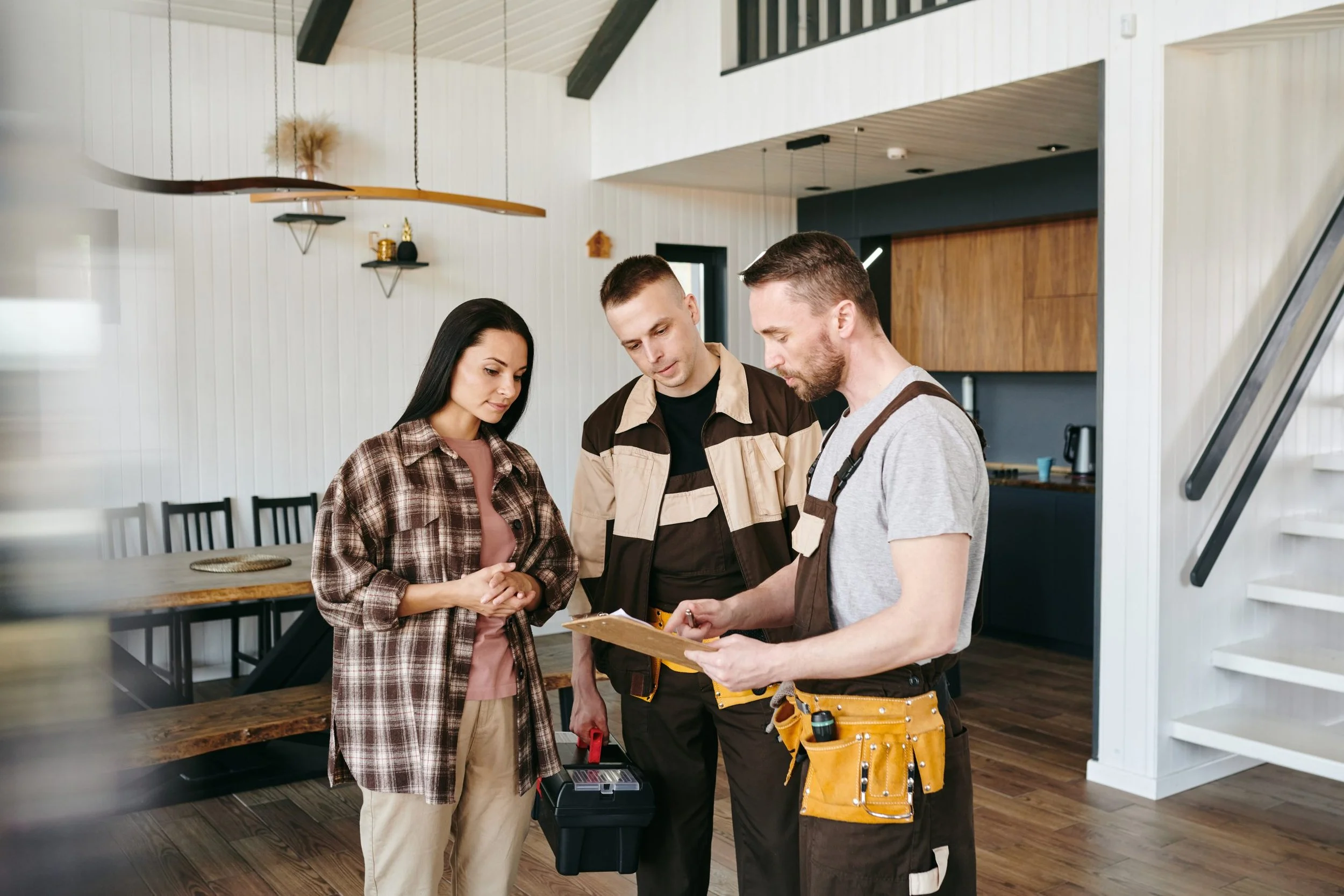 A couple discusses with a handyman in a modern kitchen, who is holding a clipboard and wearing a tool belt.