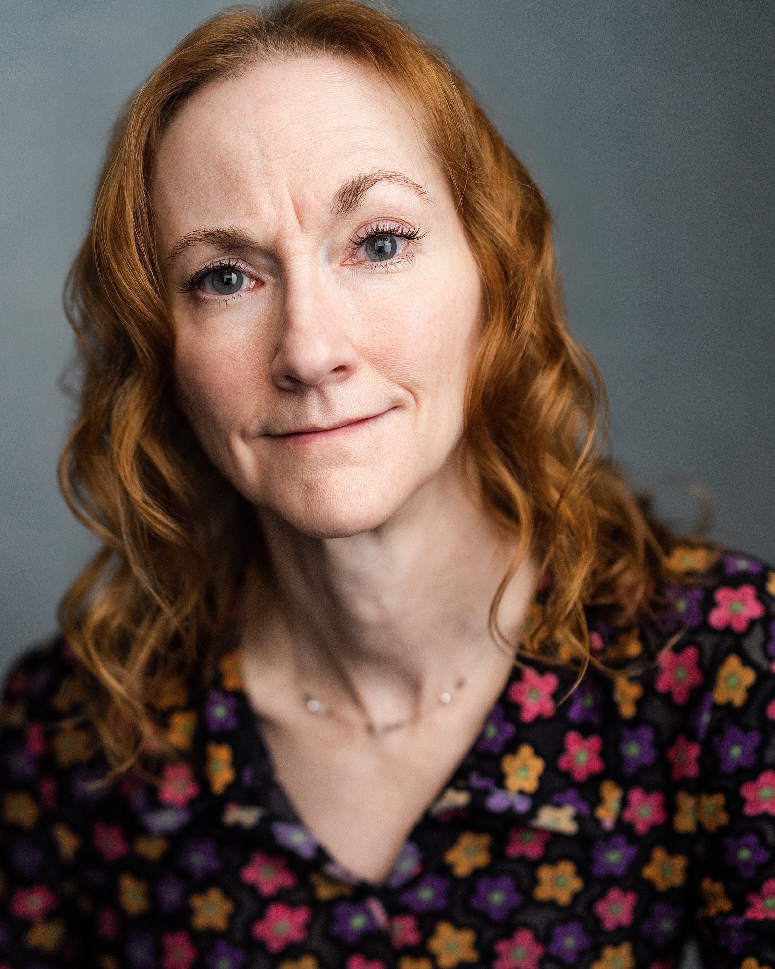 Redheaded woman with raised eyebrow and smirk. She wears a flowered v-neck top.