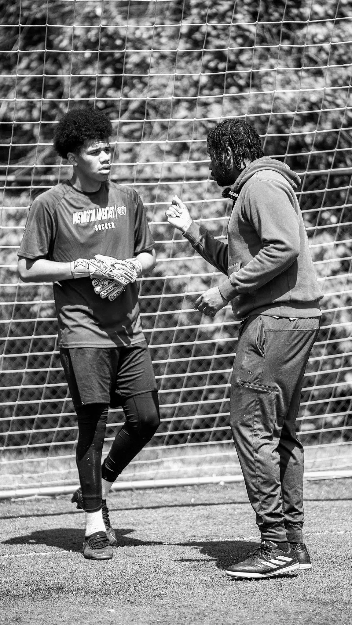 Two men, one wearing a sports jersey, gloves, and athletic pants, and the other dressed in a hoodie and joggers, are having a serious conversation near a soccer goalpost.