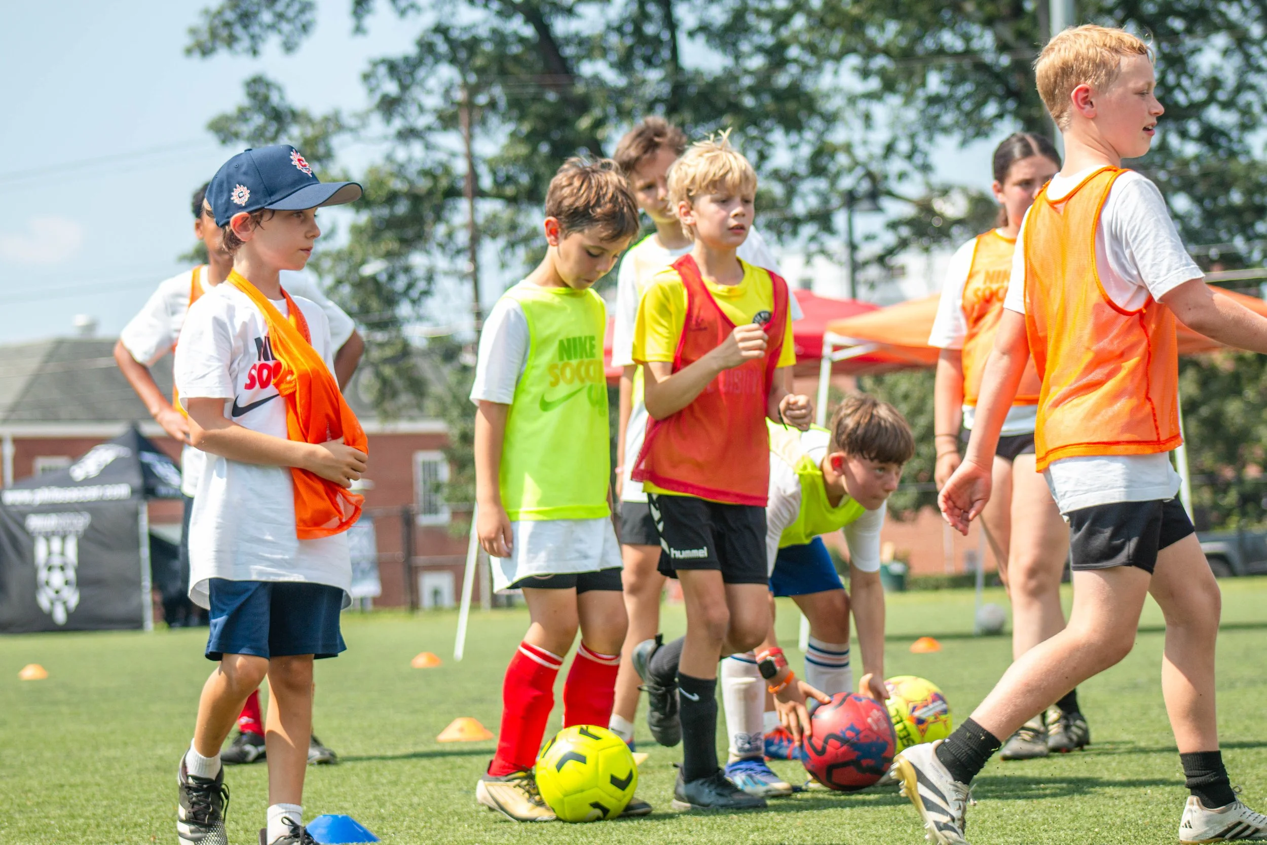 Group of young girls piled on top of each other happily on a soccer field near a goalpost, with a soccer ball nearby.