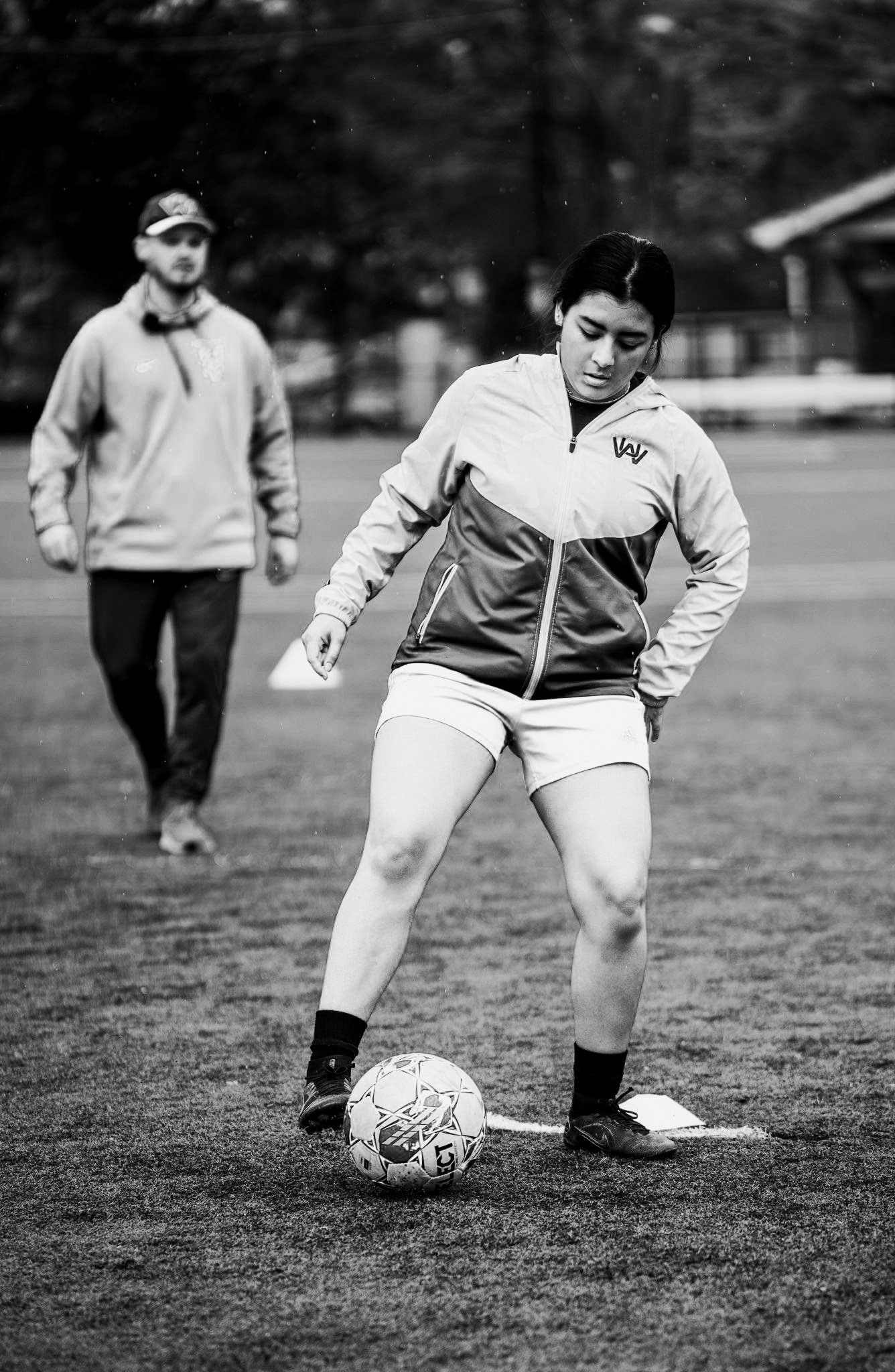 A young woman in sportswear playing soccer outdoors, with a man in a hoodie walking behind her on a grassy field.