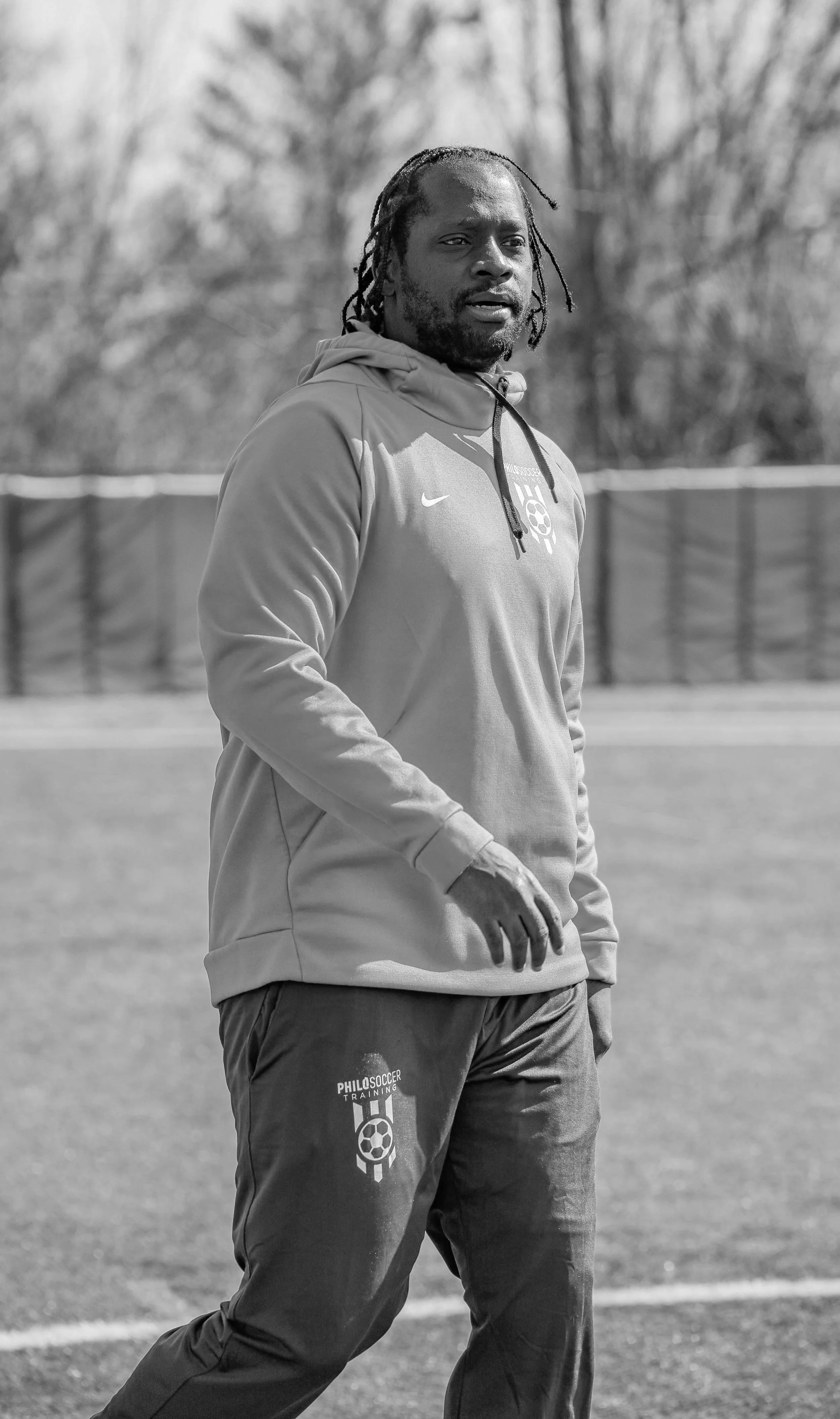 A Soccer Coach with dreadlocks wearing a hoodie and sweatpants stands on a sports field.