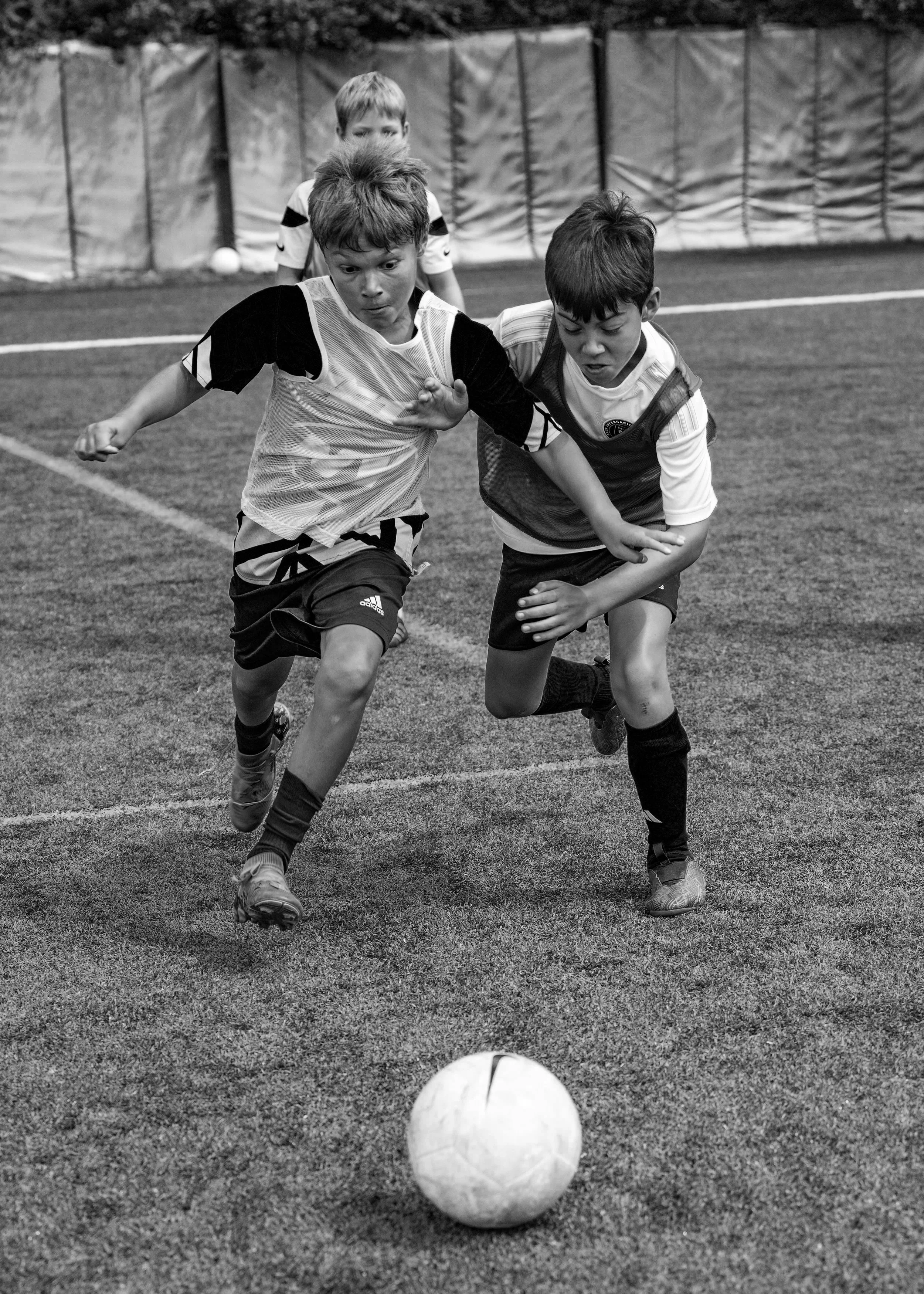 Two children playing soccer indoors, chasing the ball, with a third child watching in the background.