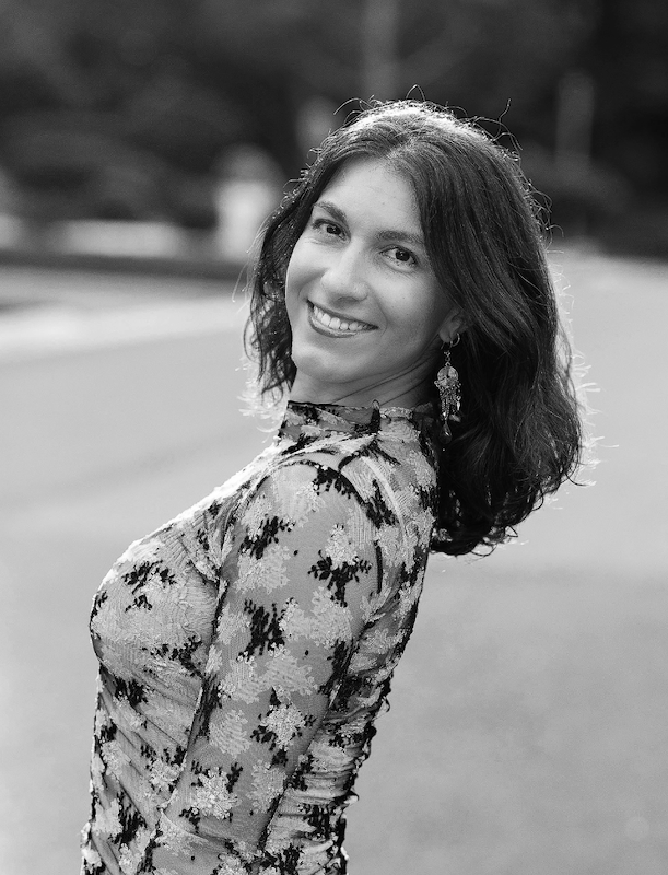 A smiling woman with shoulder-length wavy hair wearing a patterned dress standing outdoors.