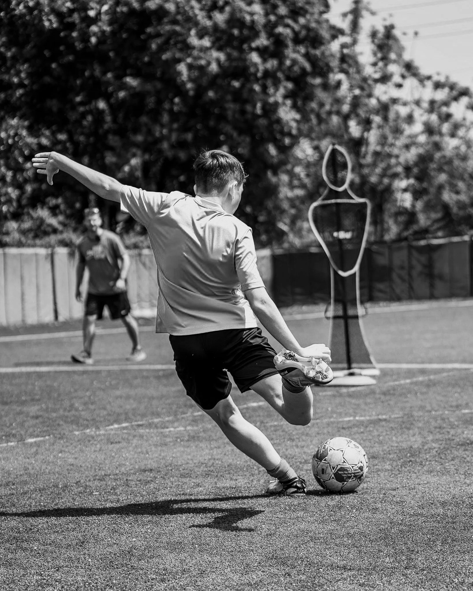 A boy in a t-shirt and shorts kicking a soccer ball on a field during outdoor practice or game, with a coach or teammate in the background, and a training dummy in the distance.