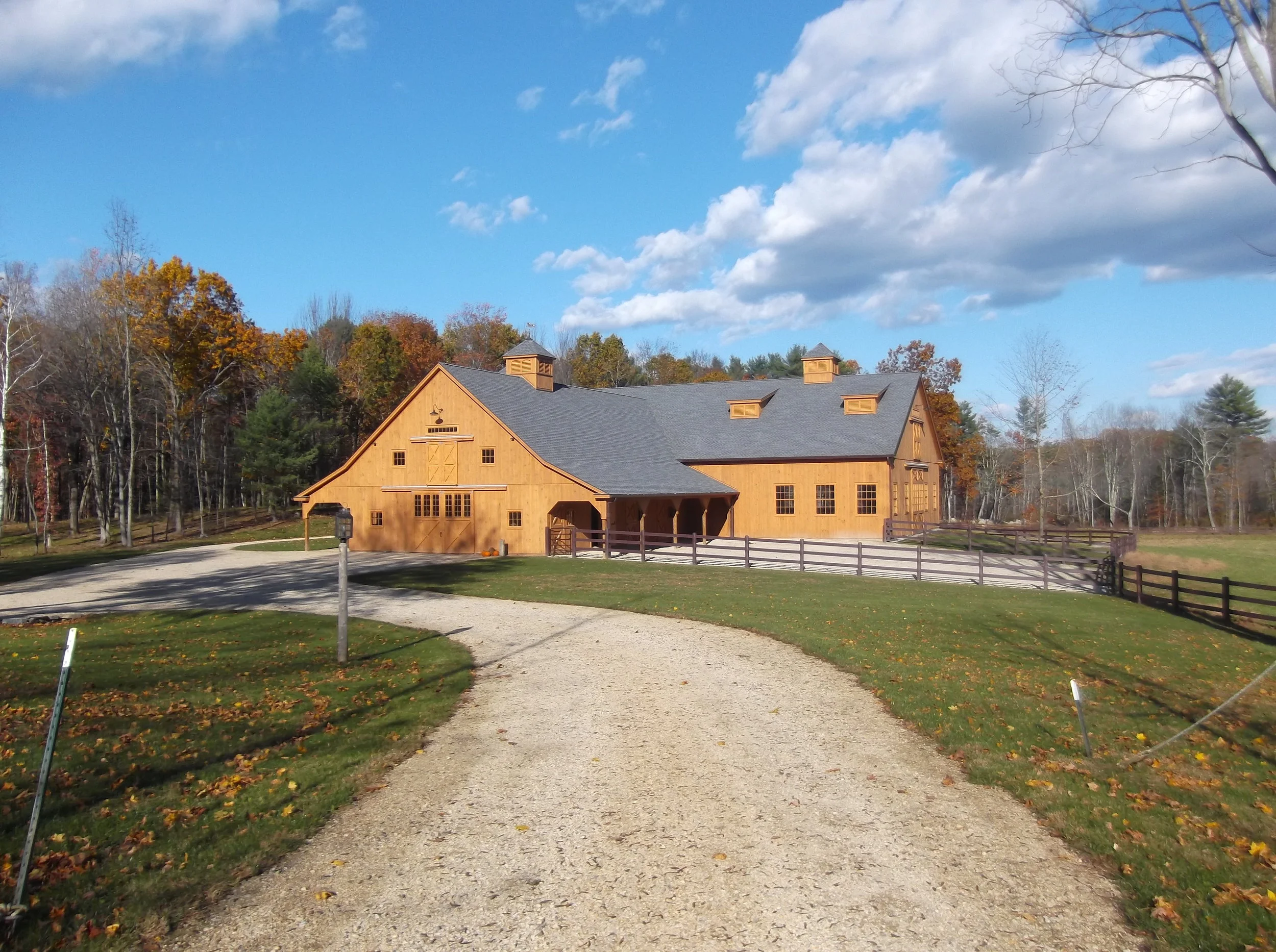  The image shows a large, wooden barn with a gray roof. The barn has two cupolas on the roof. It is surrounded by trees and a fence. There is a long driveway leading up to the barn. The barn is located in a rural setting. 