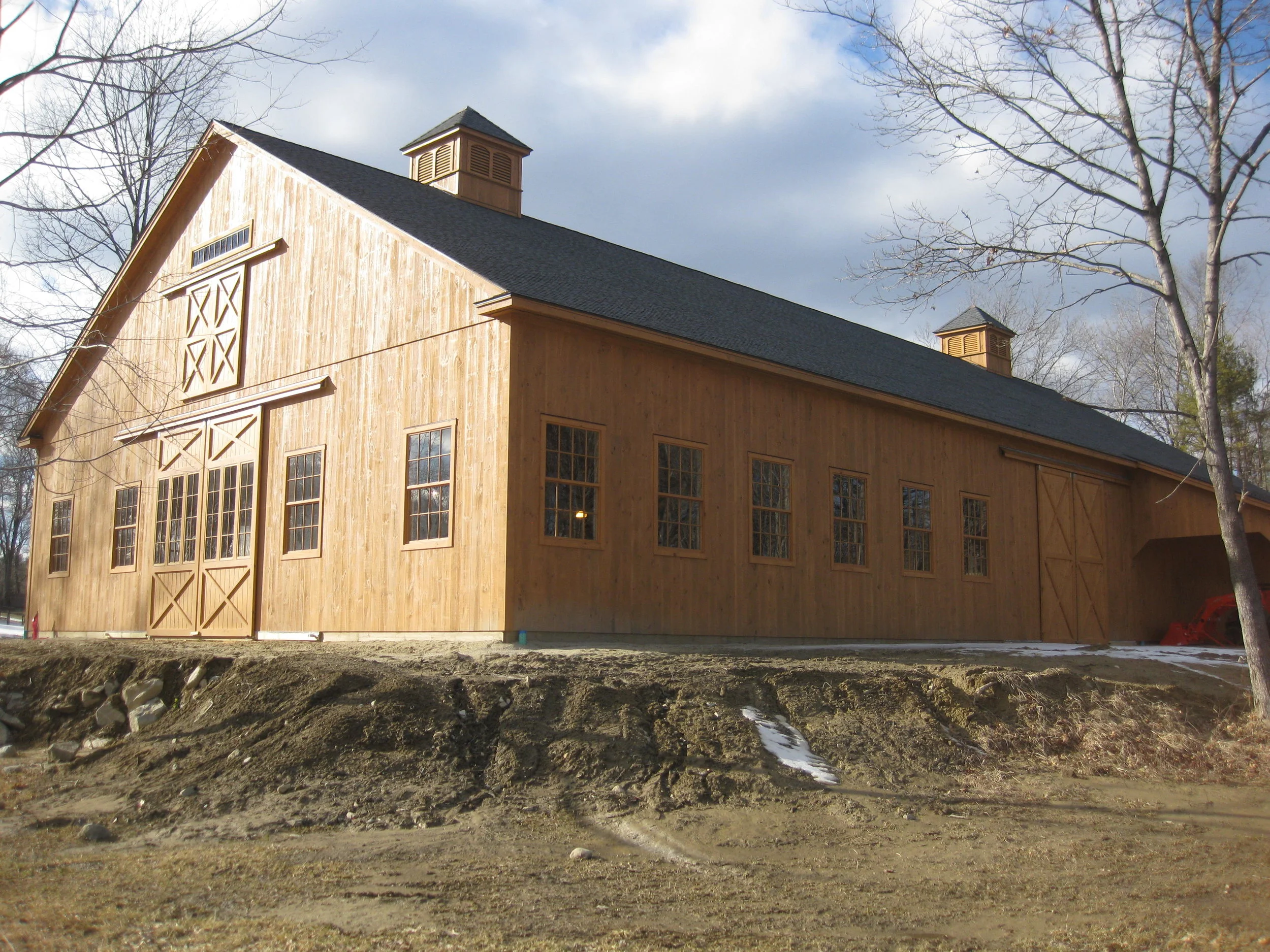  The image shows a large wooden barn. The barn is painted a light brown color and has windows. There are two large doors on the front of the barn and there are windows on both sides. The barn is located in a field and there are trees and bushes in th