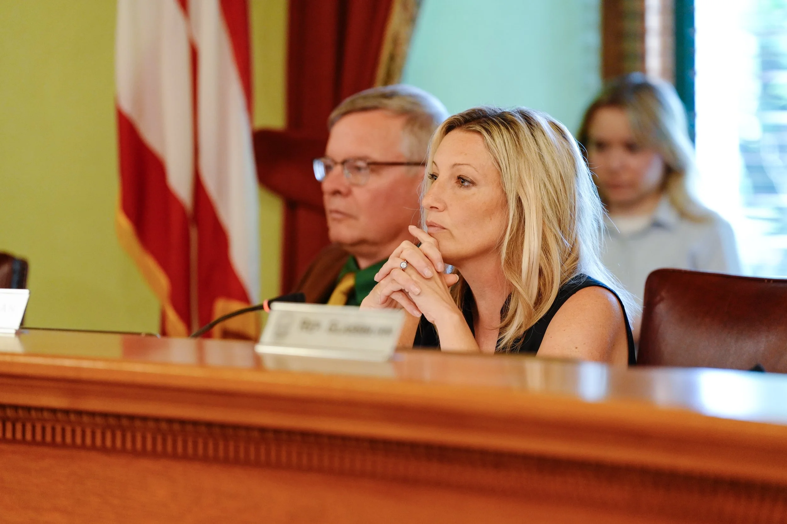 Several people seated at a meeting or conference, one woman in the foreground with blonde hair listening attentively. Background includes an American flag and curtains.