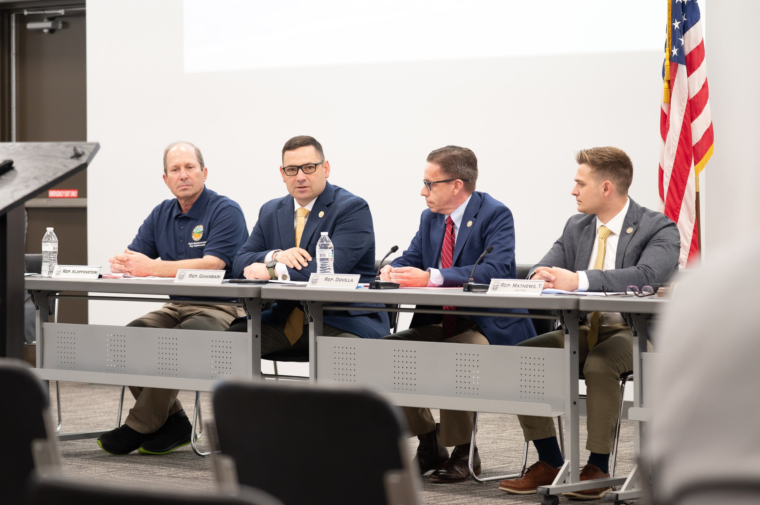Four men sitting at a conference table with water bottles, microphones, and nameplates in front of them, in a room with an American flag and a blank white wall.