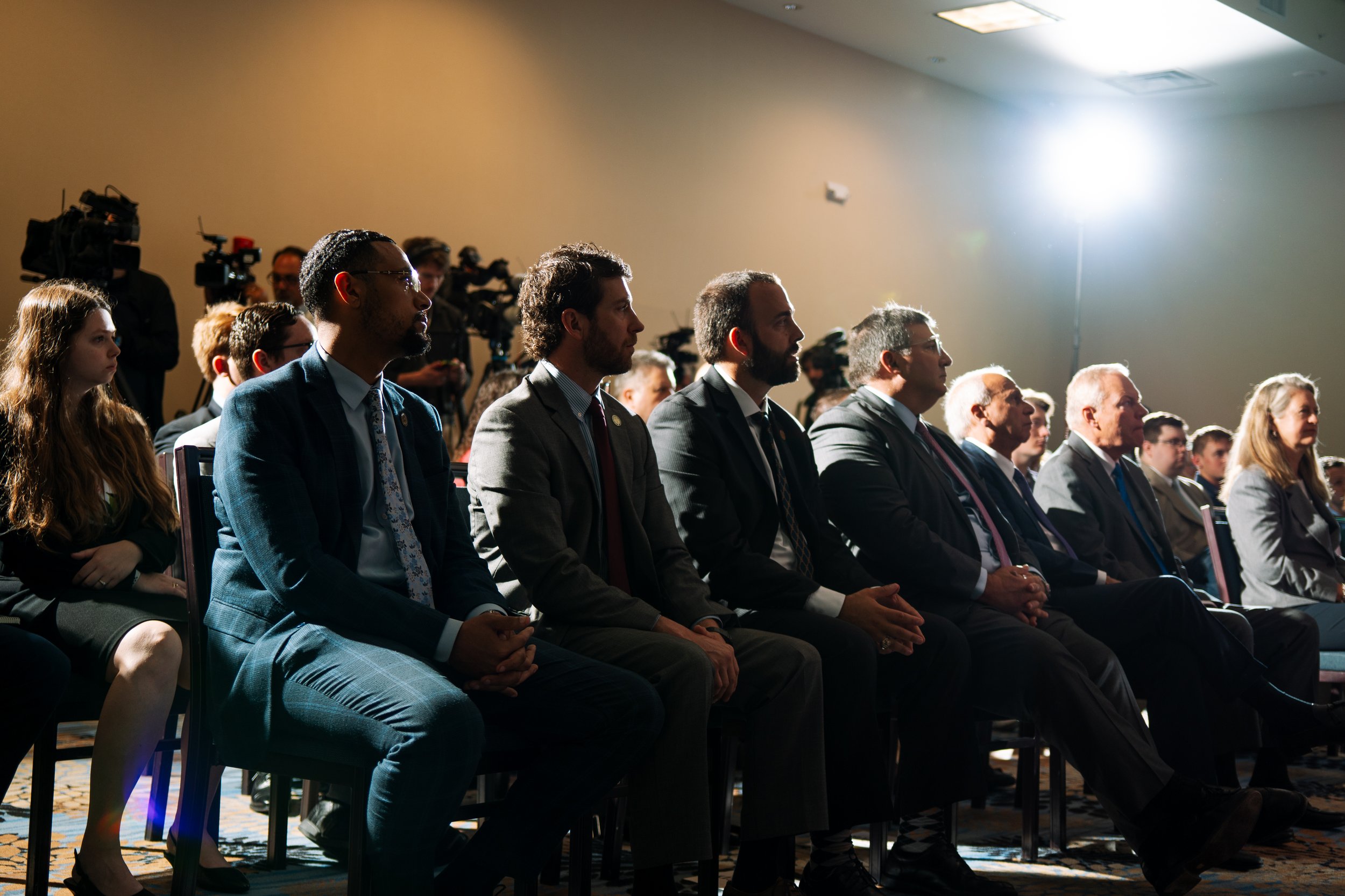 A group of people attending a formal event, sitting in rows of chairs during the day. They are dressed in suits and business attire, facing forward, with camera crews and journalists in the background.