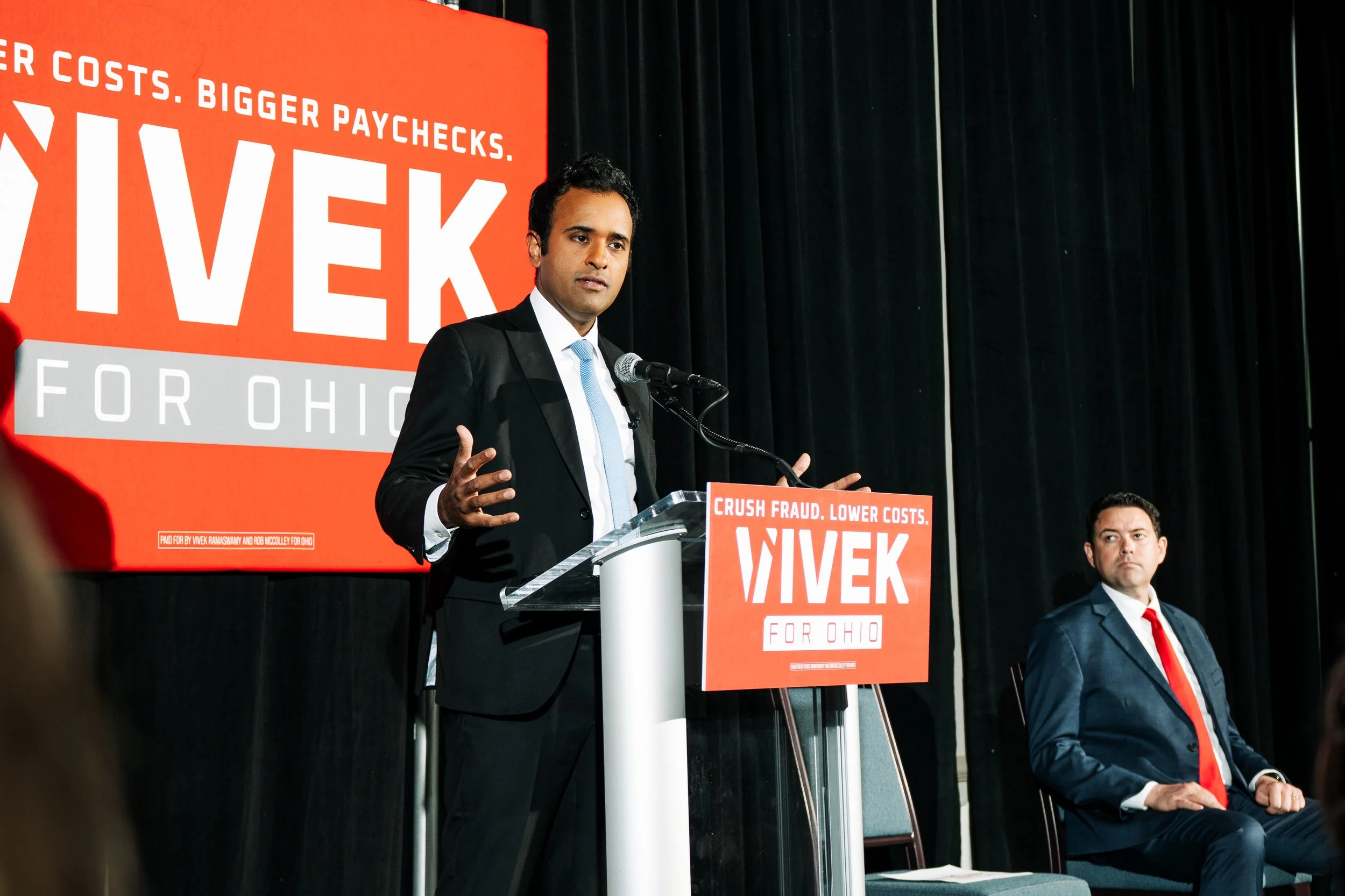 A man in a black suit and light blue tie with short black hair speaking at a podium during a live event, with a large red banner behind him displaying white text that reads "VIVEK for Ohio" and additional smaller text. Another man in a dark suit, whi