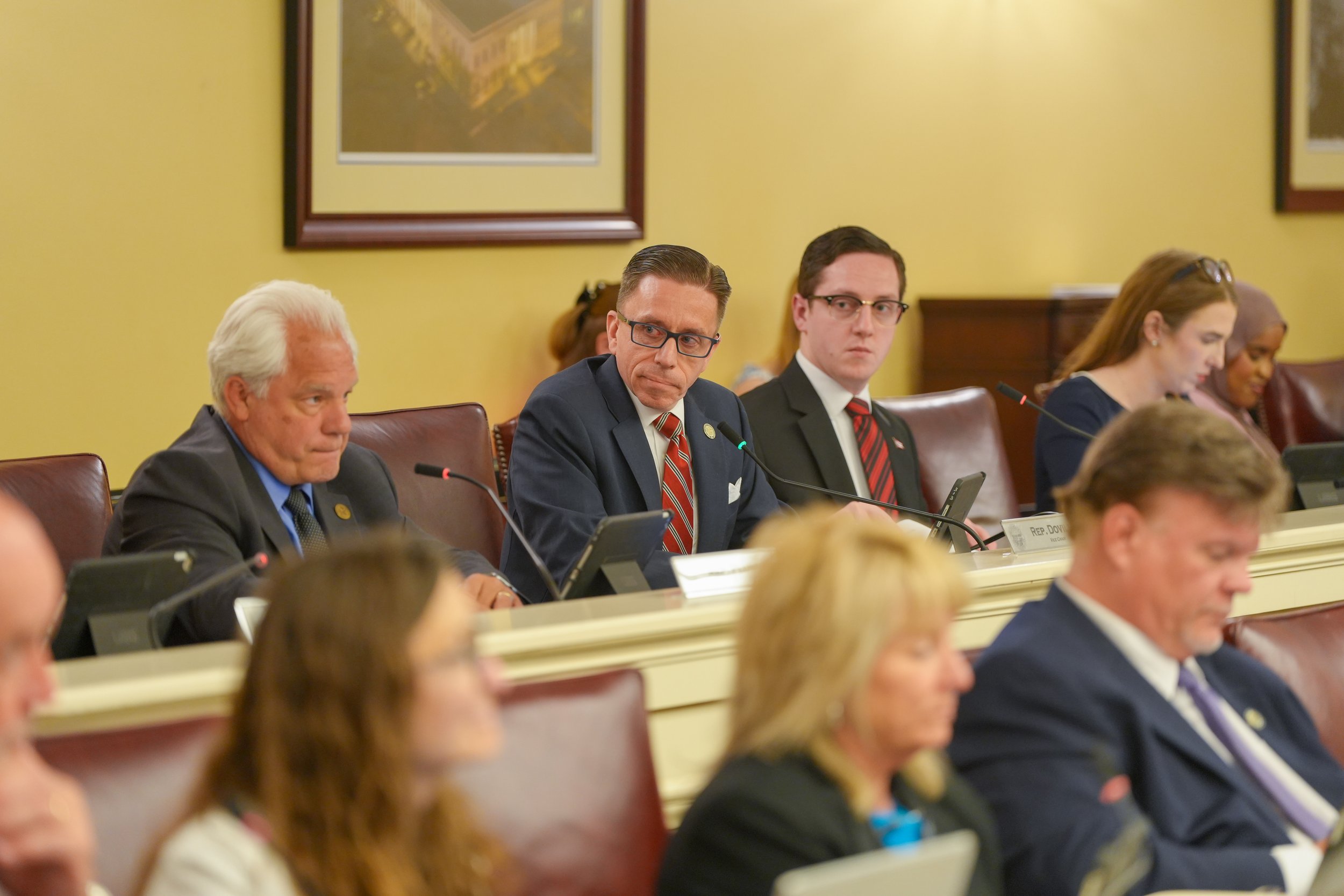 A group of people sitting at a long table in a formal setting, including a man with white hair, two men with glasses and dark suits, and a woman with glasses and red hair, participating in a meeting or hearing in a room with yellow walls, framed artw