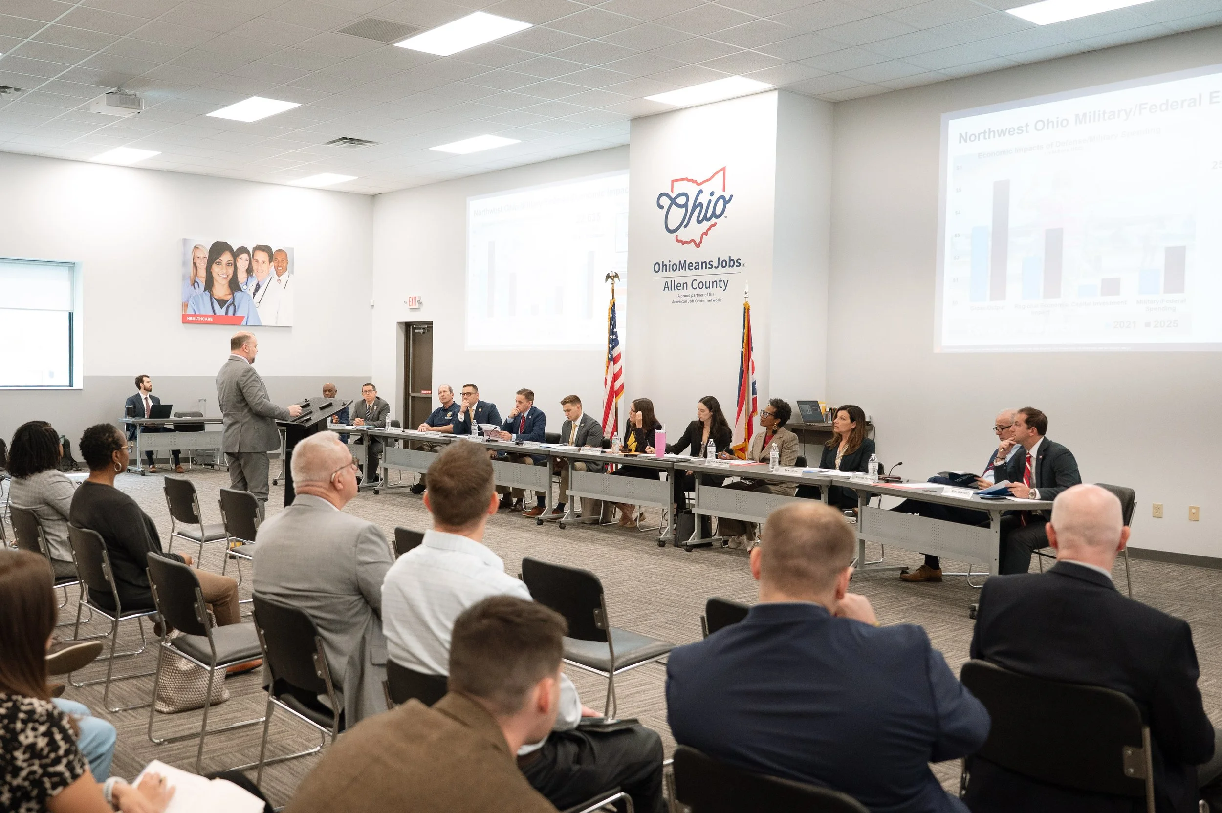 A conference room with people seated at long tables, a speaker standing at the front, presenting with slides about Ohio and military/federal topics, with flags and Ohio-themed signage.