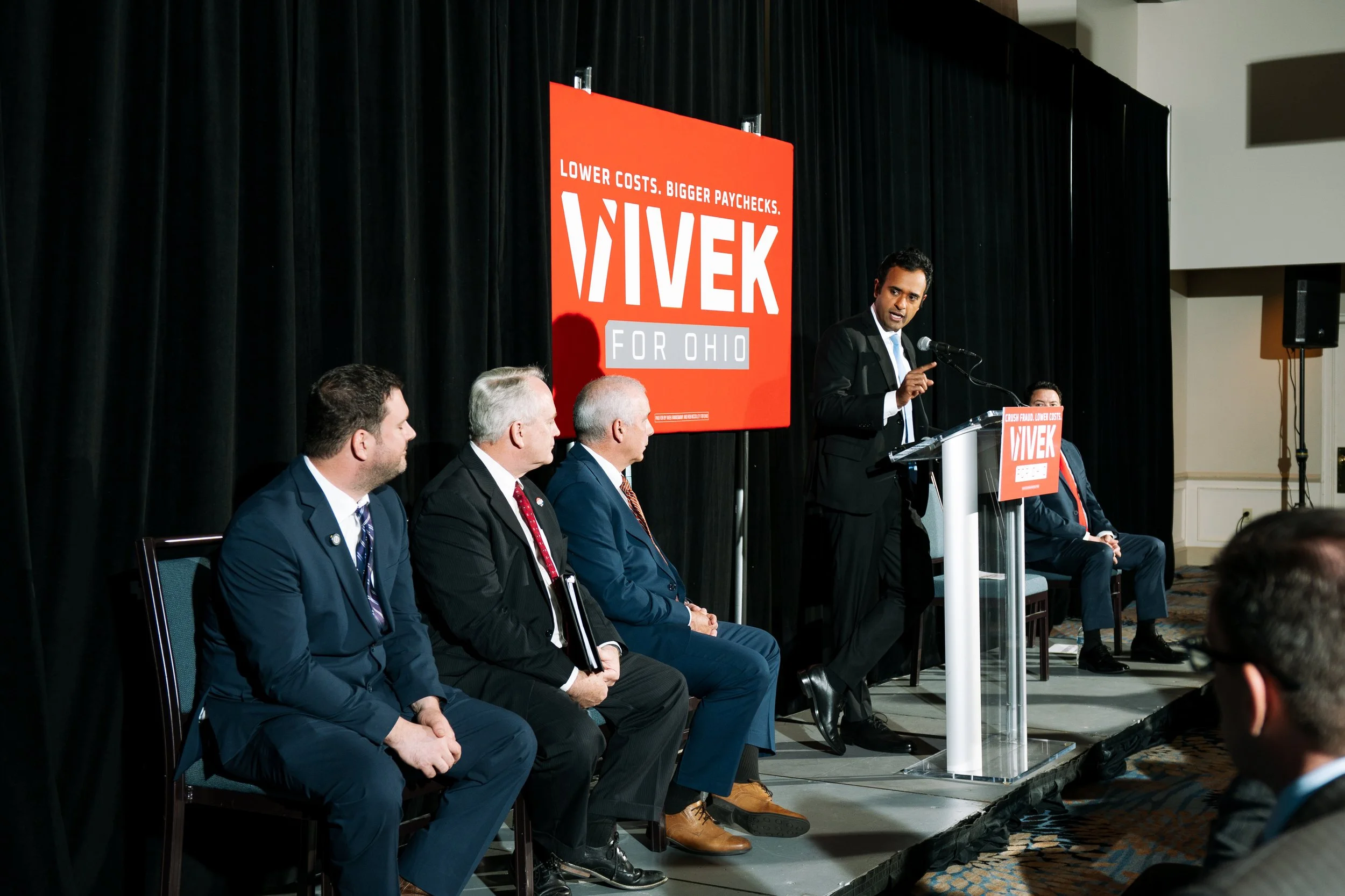 A man in a black suit speaking at a podium during a political event. Several men in suits are seated on stage. Behind the stage is a large red sign with white and gray text that reads "LOWER COSTS. BIGGER PAYCHECKS! VIVEK FOR OHIO."