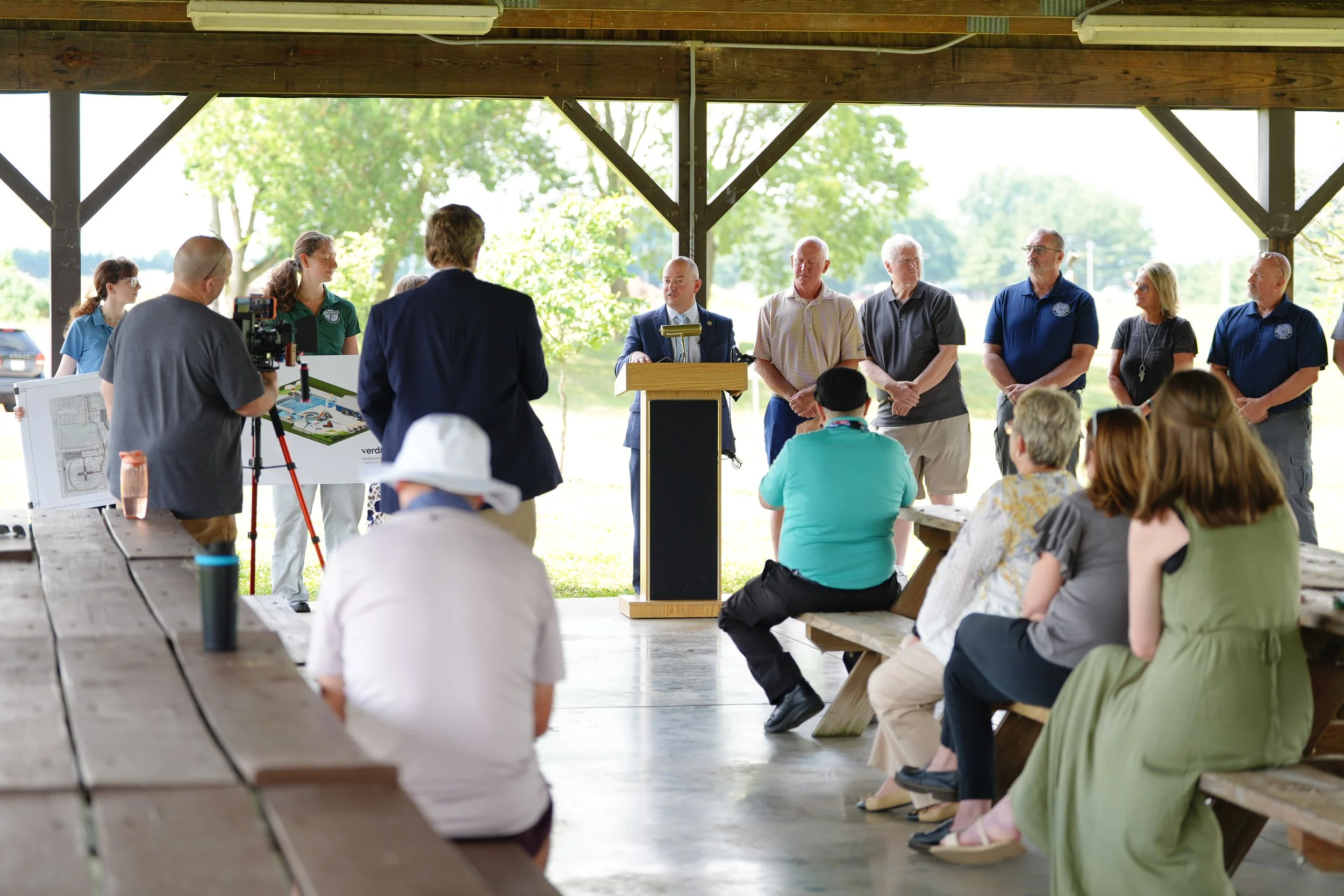 An outdoor meeting with several people standing and sitting under a pavilion, with some cameras and presentation materials visible.