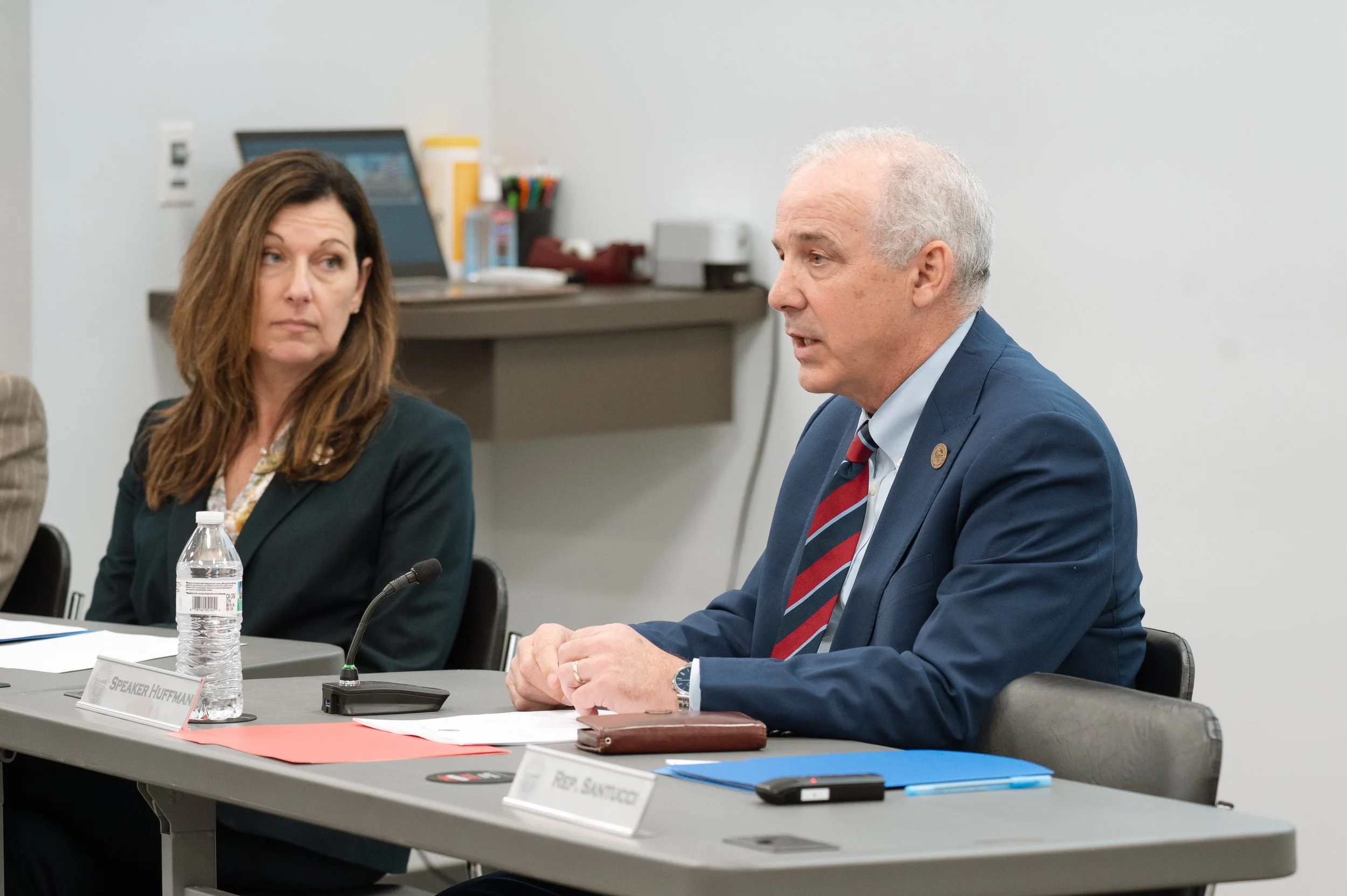 A man in a blue suit with a striped tie is speaking at a meeting, sitting at a conference table with a woman next to him. There are documents, a water bottle, and microphones on the table.