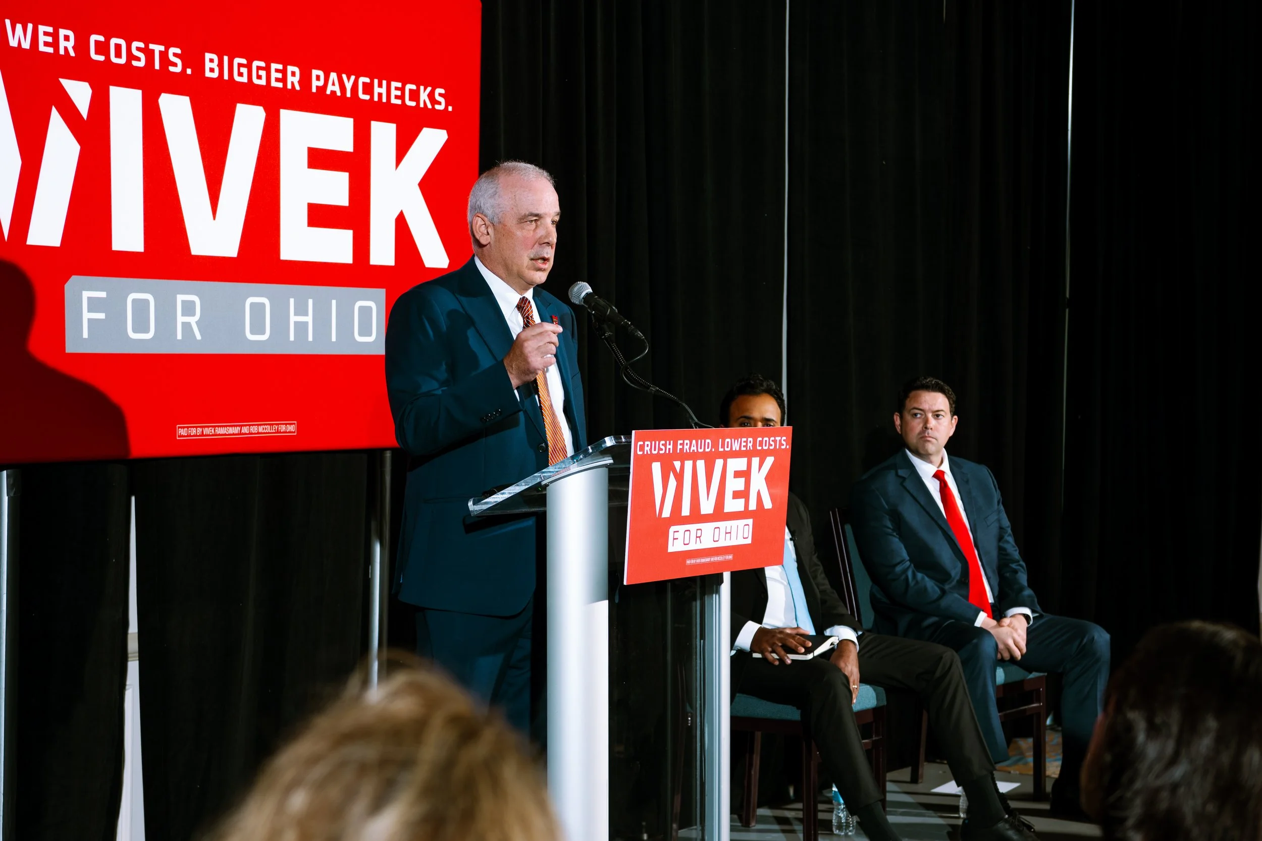A man in a suit speaking at a podium with a microphone during a political event with a red campaign sign for 'VIVEK for Ohio' behind him. Two other men are seated nearby on stage, one in a suit with a red tie and the other in a black suit with an ope