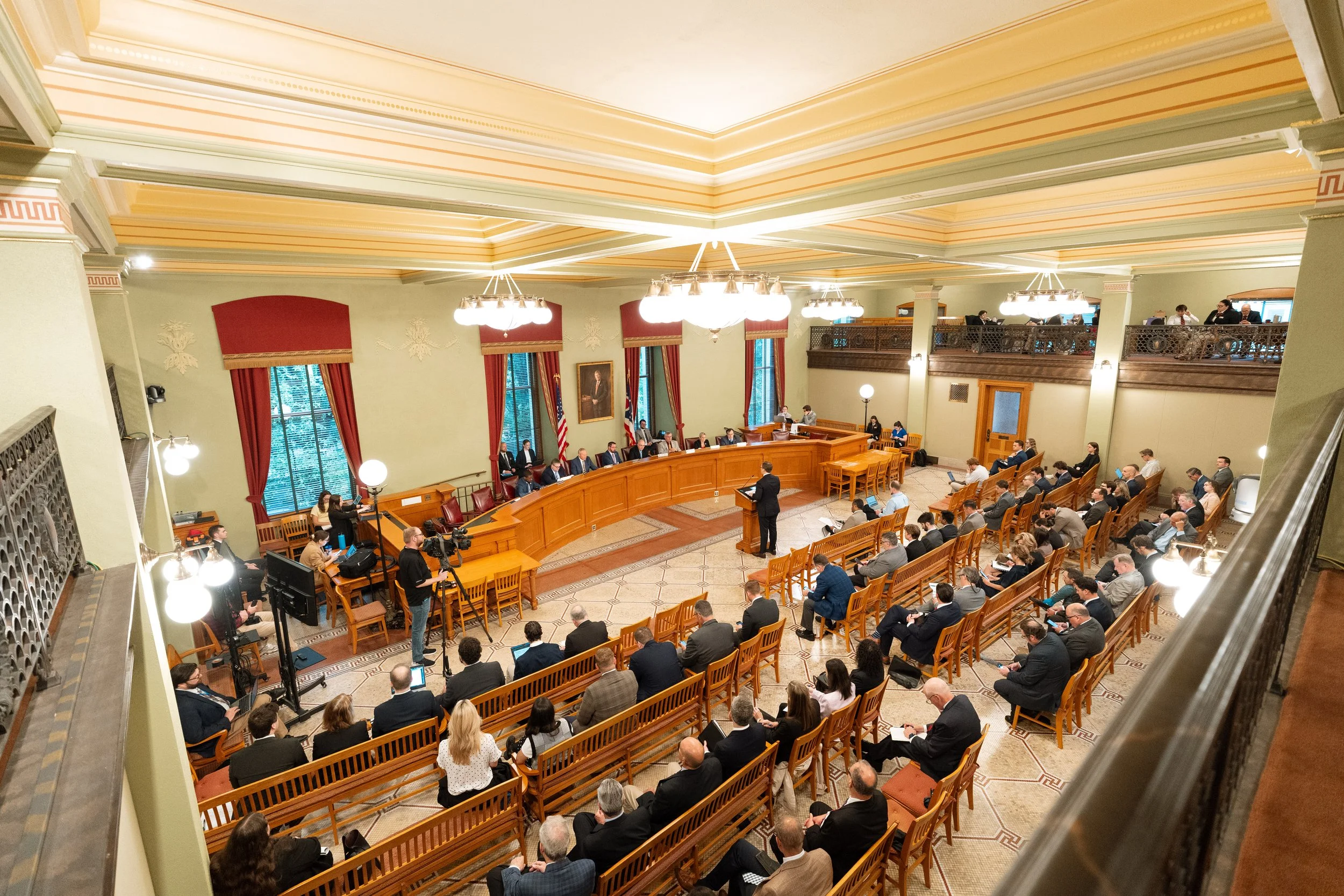 A formal government meeting inside a large, ornate room with high ceilings and chandeliers. People are seated at a large, curved table and on chairs in rows, with some standing and speaking. There are flags, a portrait, and multiple people using lapt