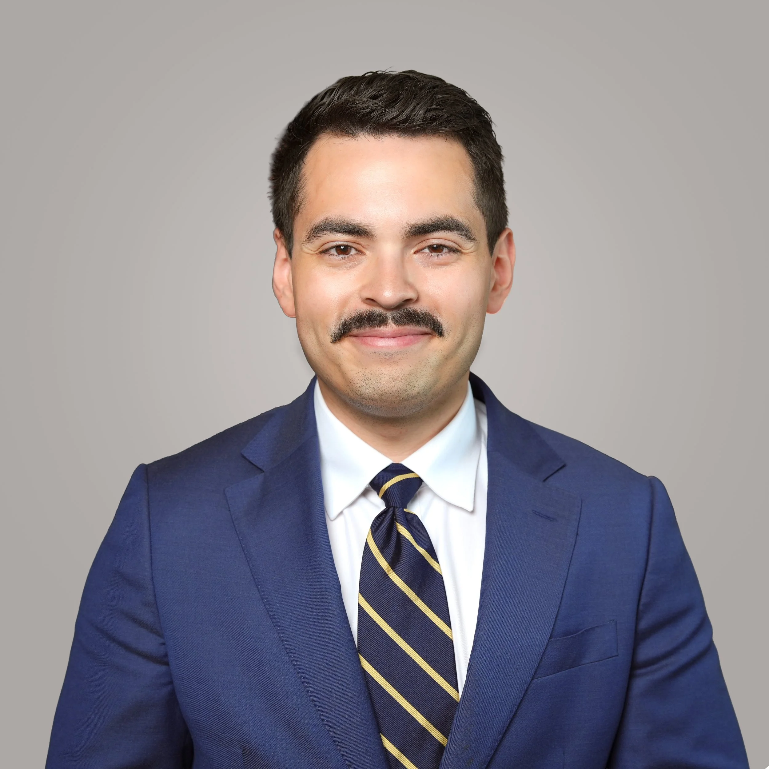 A man wearing a navy blue suit, a white dress shirt, and a dark blue tie with yellow diagonal stripes, smiling against a plain gray background.
