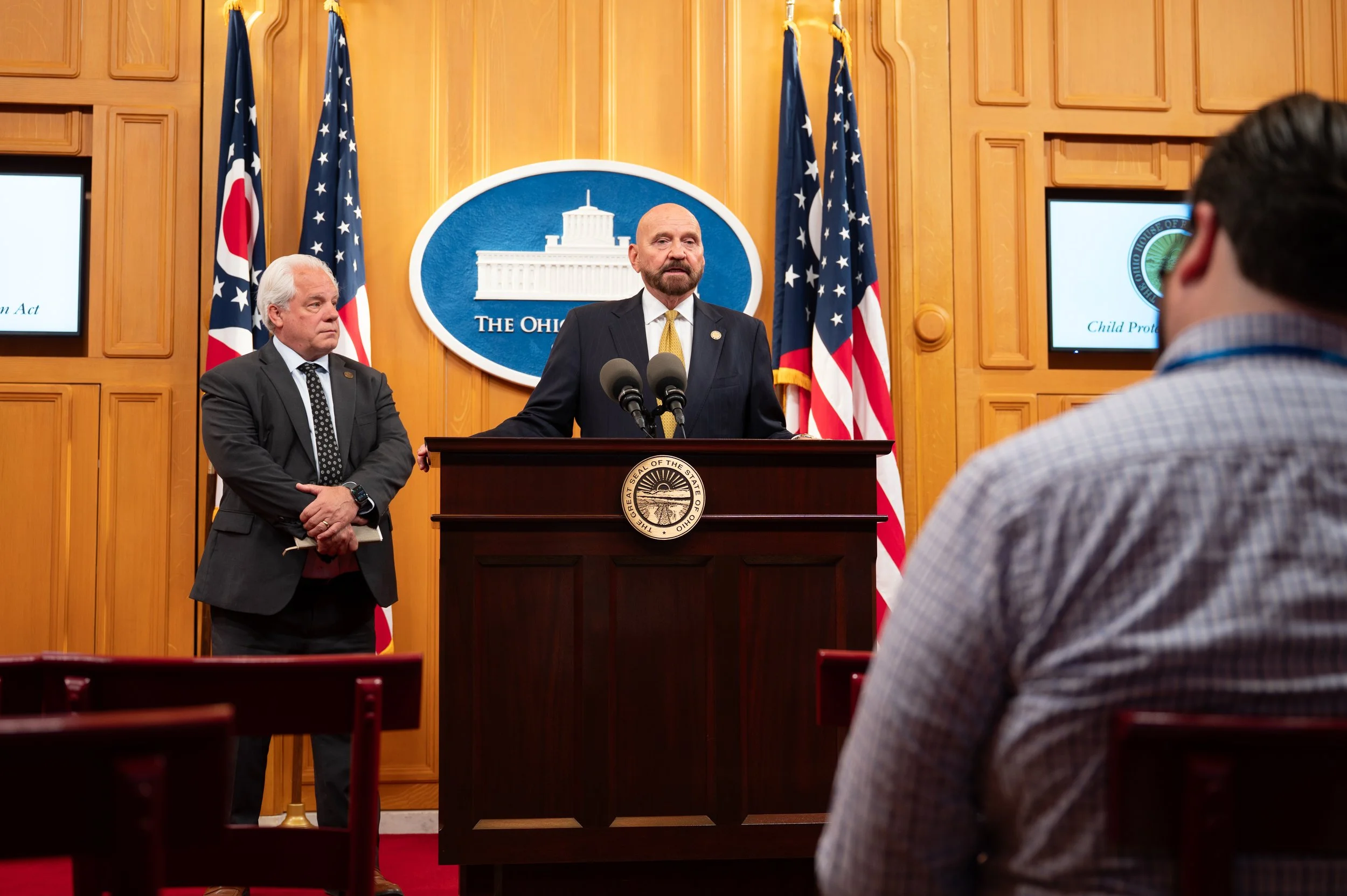 A man with a beard and suit speaking at a podium with a seal that reads 'The Ohio,' flanked by flags of Ohio and the United States, with a man in a suit standing next to him. An audience member is seen in the foreground.