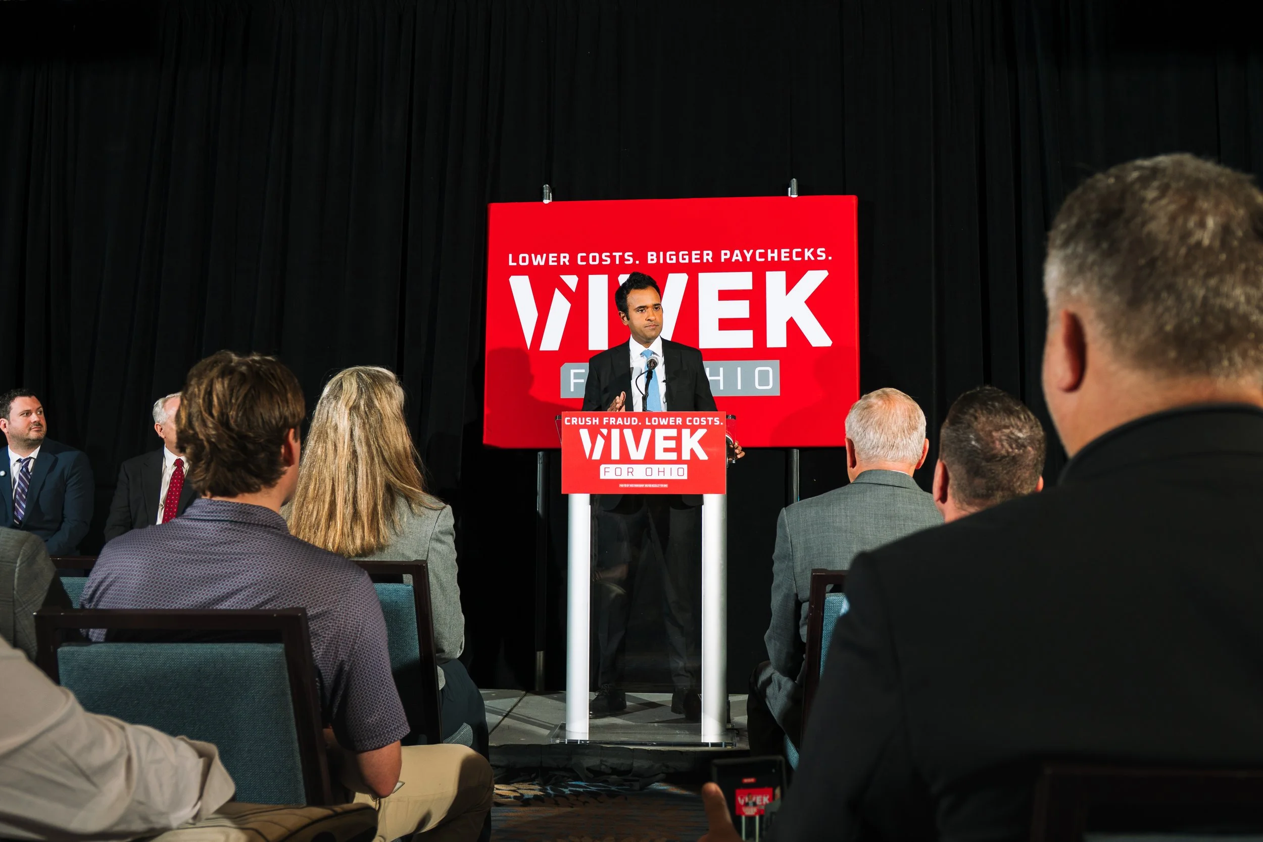 A man in a suit and tie giving a speech at a podium with a red and white campaign sign that reads "VIVEK FOR OHIO" and slogans "Lower costs. Bigger paychecks." and "Crush fraud. Lower costs." in front of a black curtain, with several people seated, s
