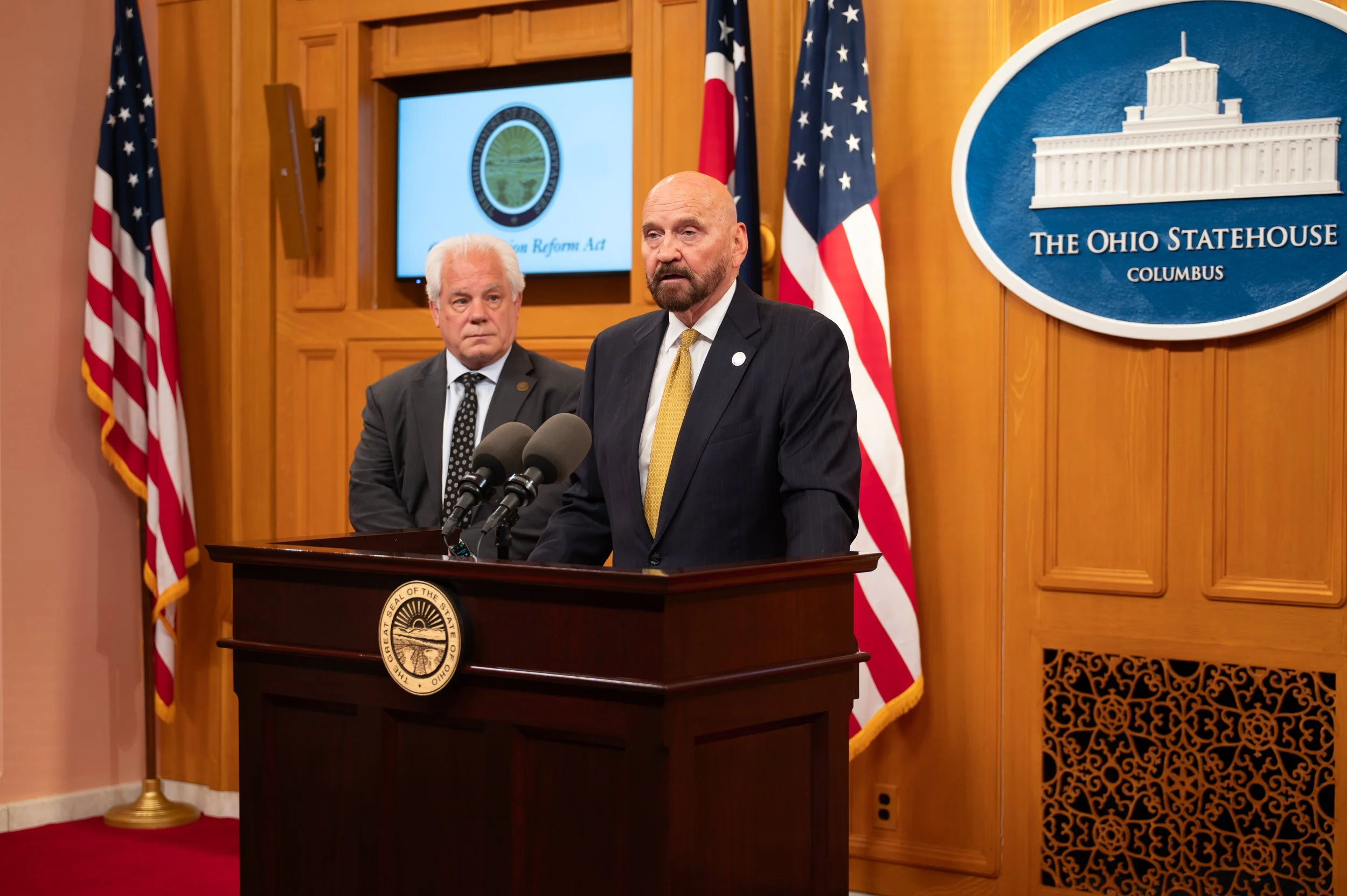 Two men in suits standing at a podium during a press conference at the Ohio Statehouse. American flags and a digital screen displaying the Ohio Legislature seal are visible in the background.