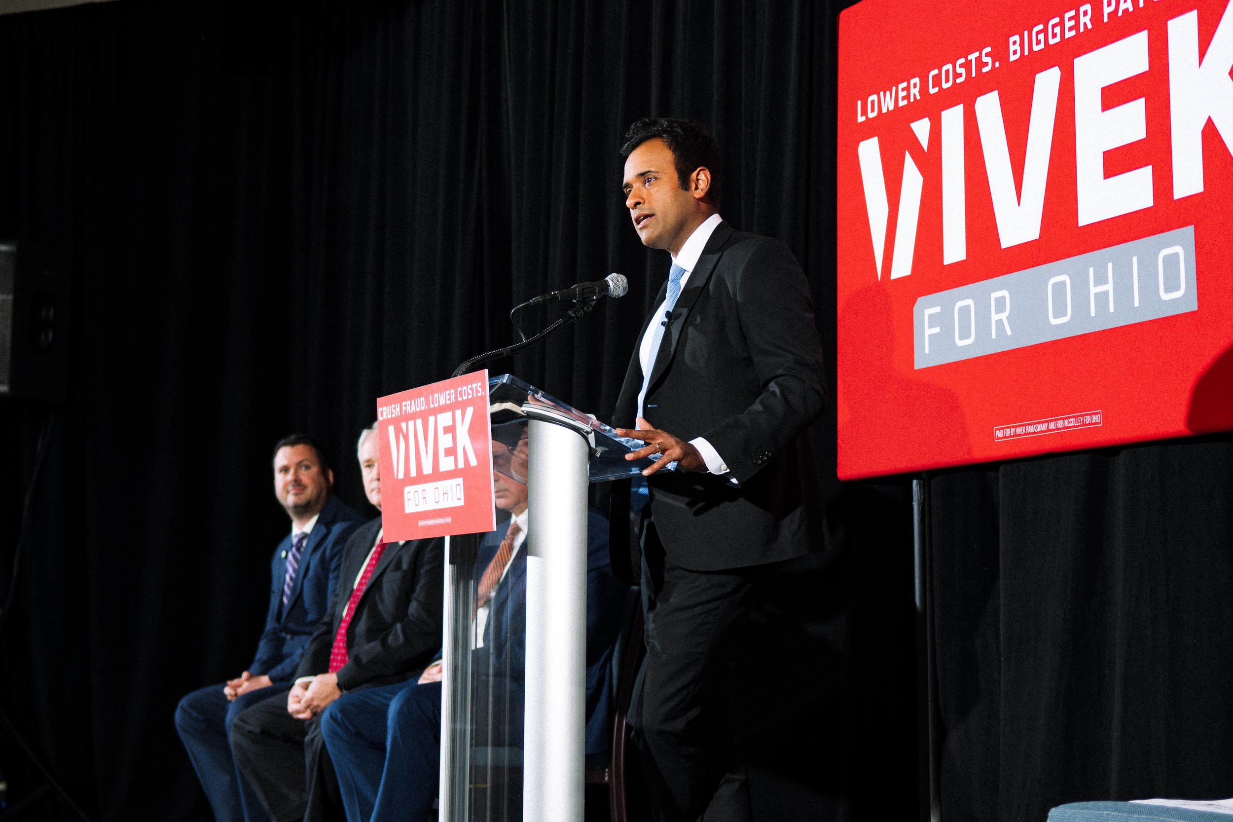 A man in a suit speaking at a podium during a political event, with a large red banner behind him displaying the words 'Lower costs. Bigger paychecks' and 'VIVEK FOR OHIO'. Several individuals are seated on stage beside him.