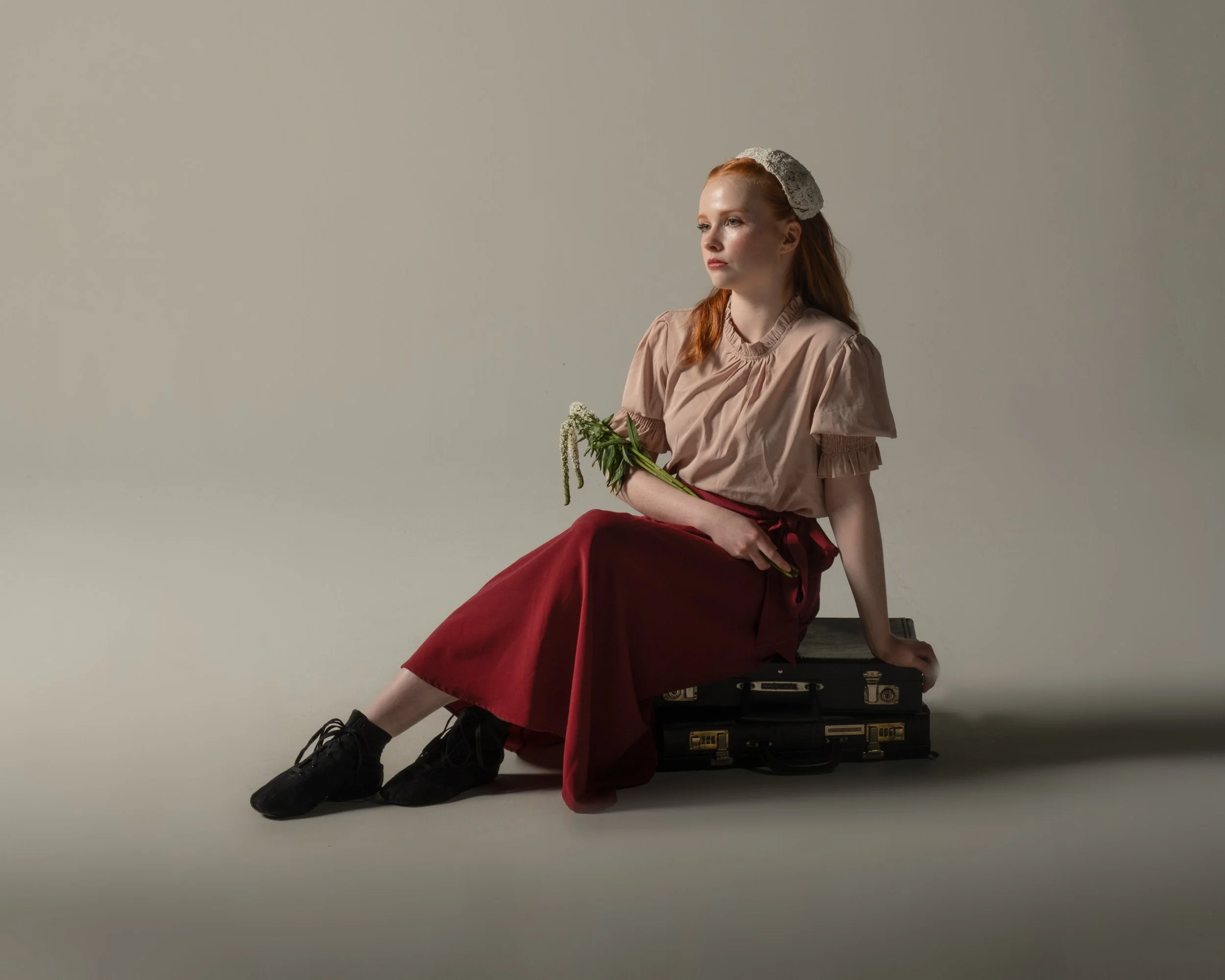 a female dancer sits on a piece of vintage luggage, holding a wilted flower and looking off into the distance with a look of melancholy on her face