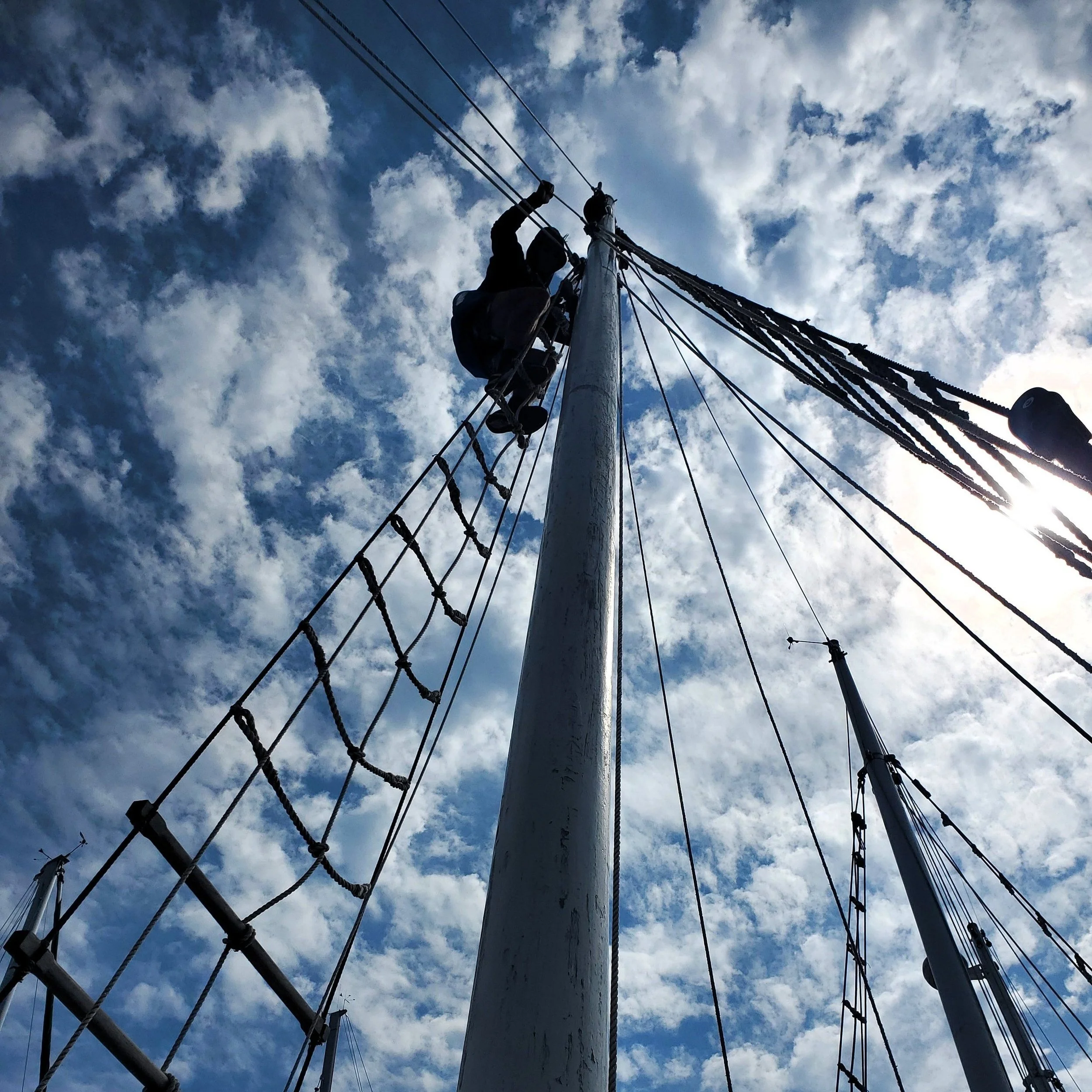 Person climbing a boat's mast against a sky with scattered clouds, viewed from below.