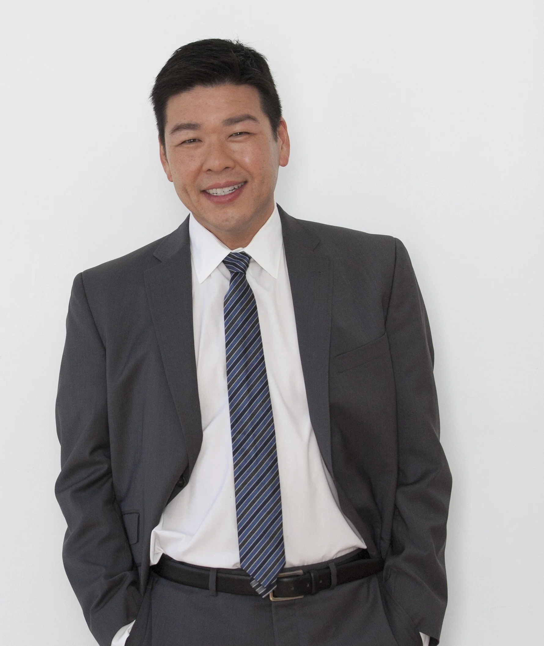 A smiling man in a dark gray business suit, white shirt, and striped tie standing against a white background.