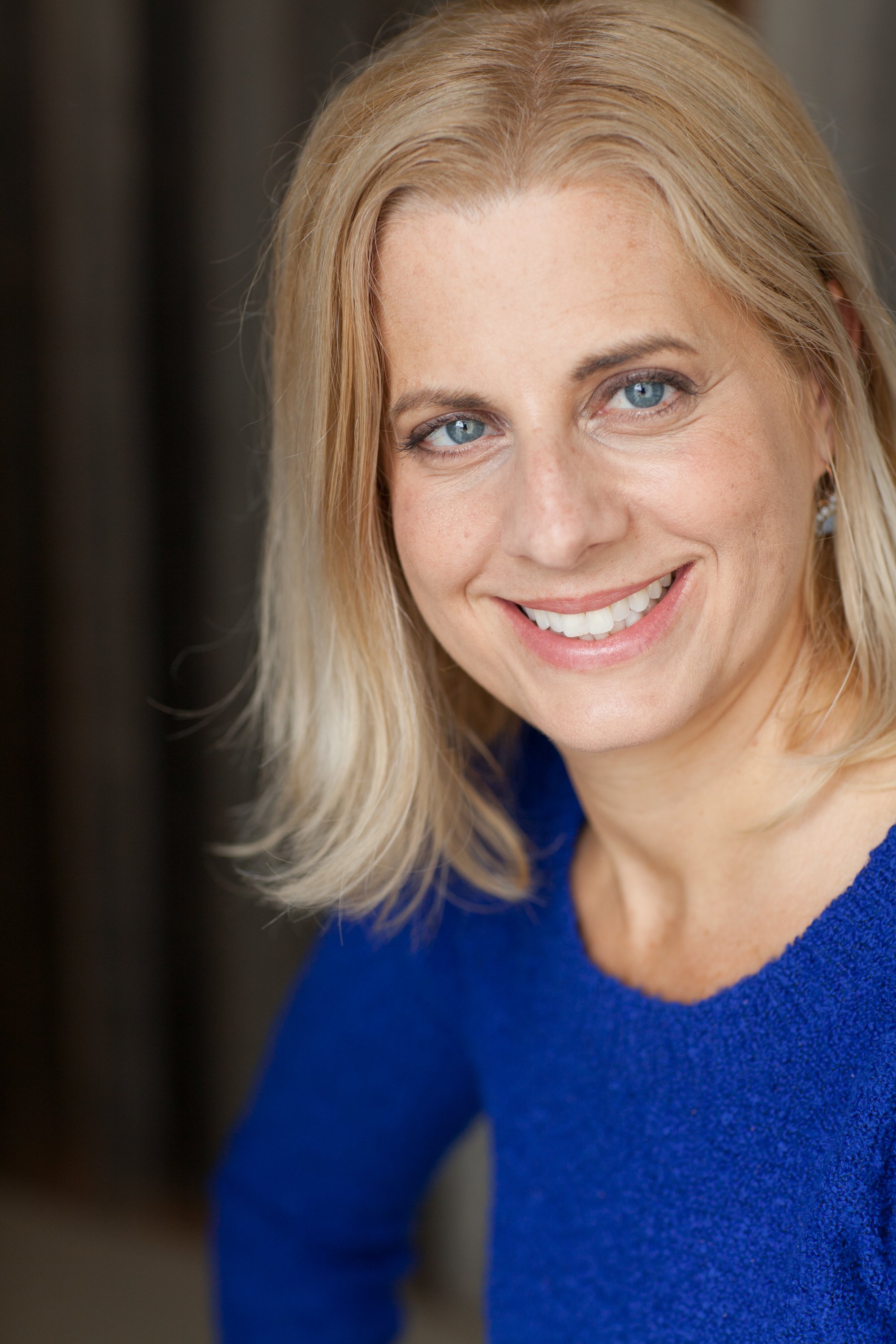 Close-up portrait of a woman with blonde hair, blue eyes, and a bright smile, wearing a blue top.