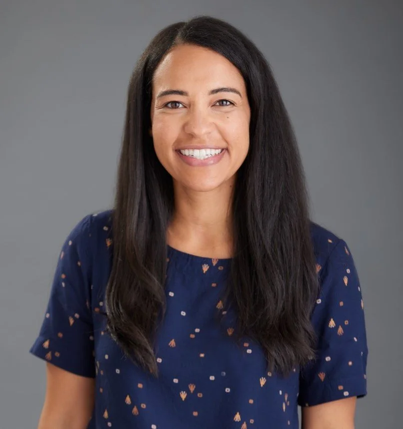 Smiling woman with long dark hair wearing a navy blue shirt with small colorful patterns against a gray background.