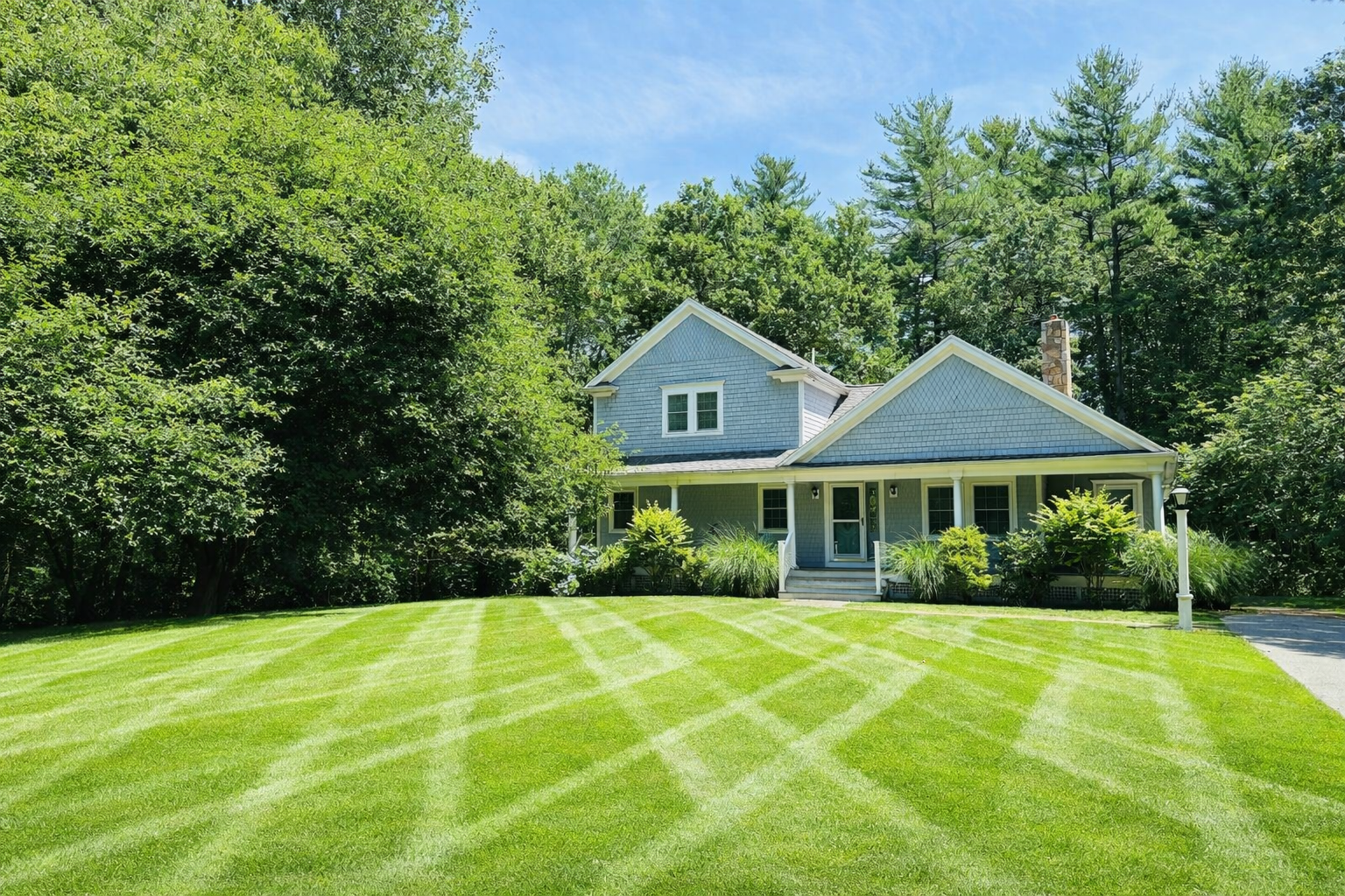 A two-story house with gray siding and white trim sits behind a neatly mowed lawn with striped grass patterns, surrounded by green trees and shrubs, under a clear blue sky.