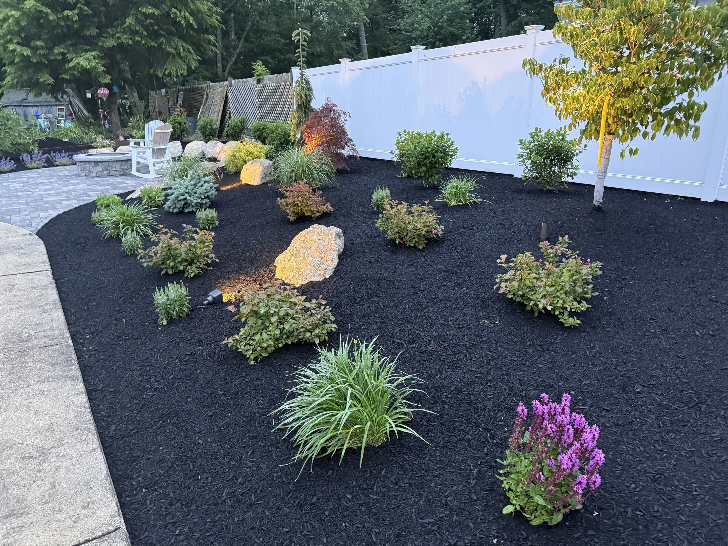 A landscaped backyard garden with black mulch, various shrubs, and small trees. There is a stone pathway with patio furniture, including a picnic table with chairs. A white privacy fence surrounds the garden, with trees and decorative elements in the background.