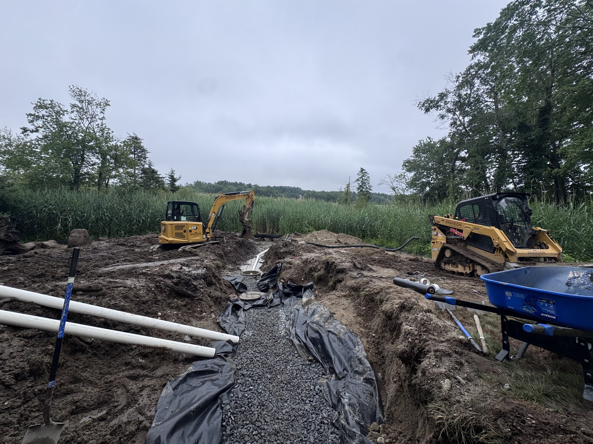 Construction site with two mini excavators working on trenching, outdoor setting with trees and cloudy sky.