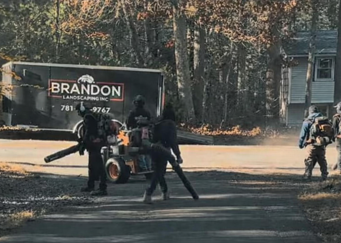 Four people in dark clothing and masks working with equipment on a street with a large Brandon Landscaping truck in the background. Two individuals are on the left, one near a small machine, and two more are on the right, one carrying a bag. The setting appears to be in a wooded area with a house visible on the right.